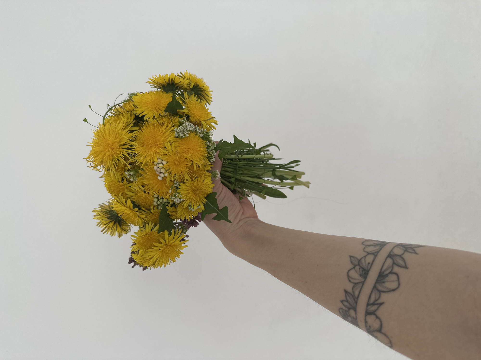 A person's forearm is held up, holding a bouquet of yellow dandelions with some small white flowers mixed in. The arm has a floral band tattoo around the upper part. The background is a plain white wall. The location in the city of Votkinsk, Russia cannot be determined from the image.