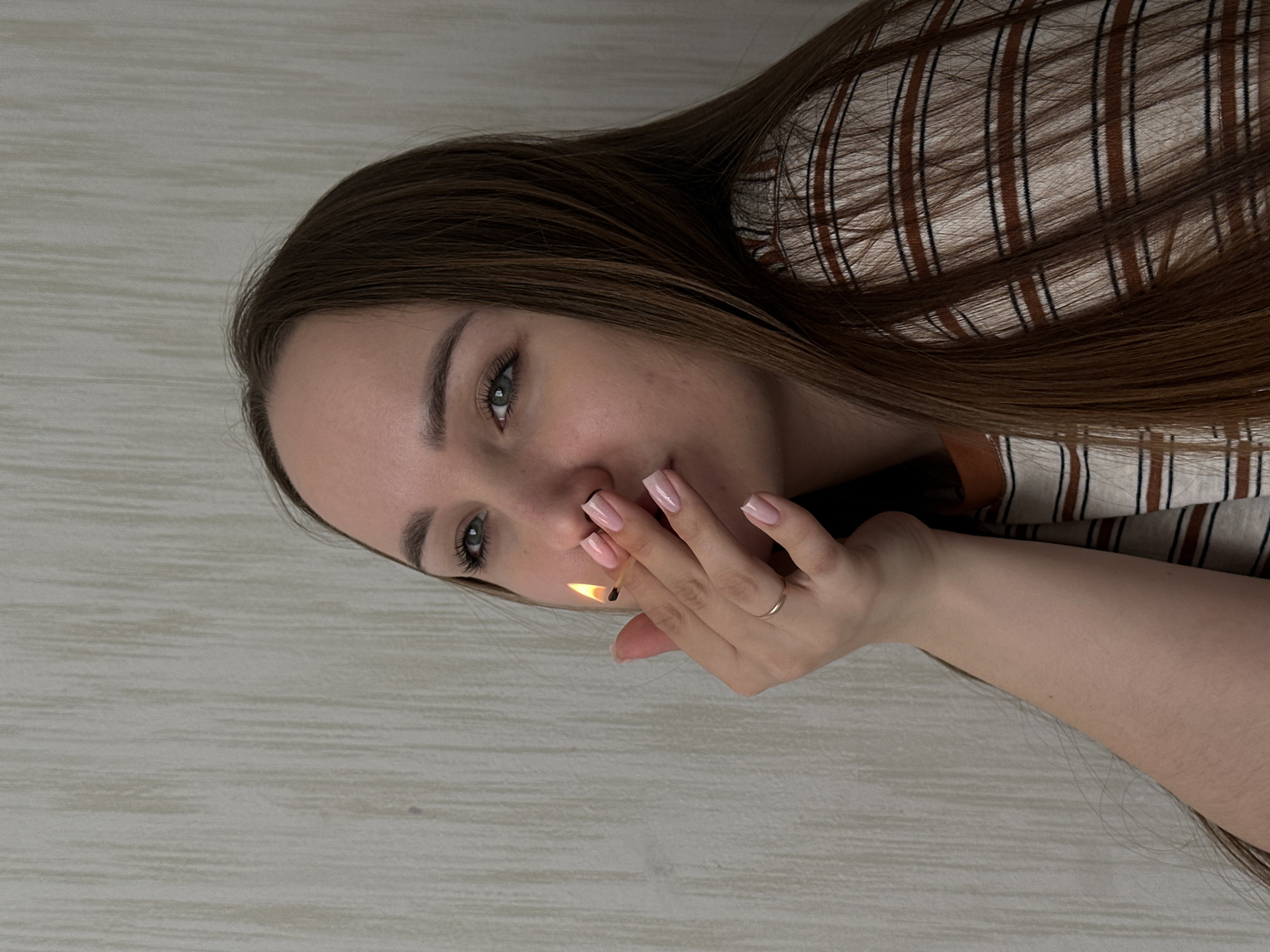 A young woman with long brown hair and light skin holds a lit match close to her face. She is wearing a striped shirt and a ring on her finger. The background is a light-colored wall. There is no verifiable information to suggest the image was taken in Chaikovsky, Russia.