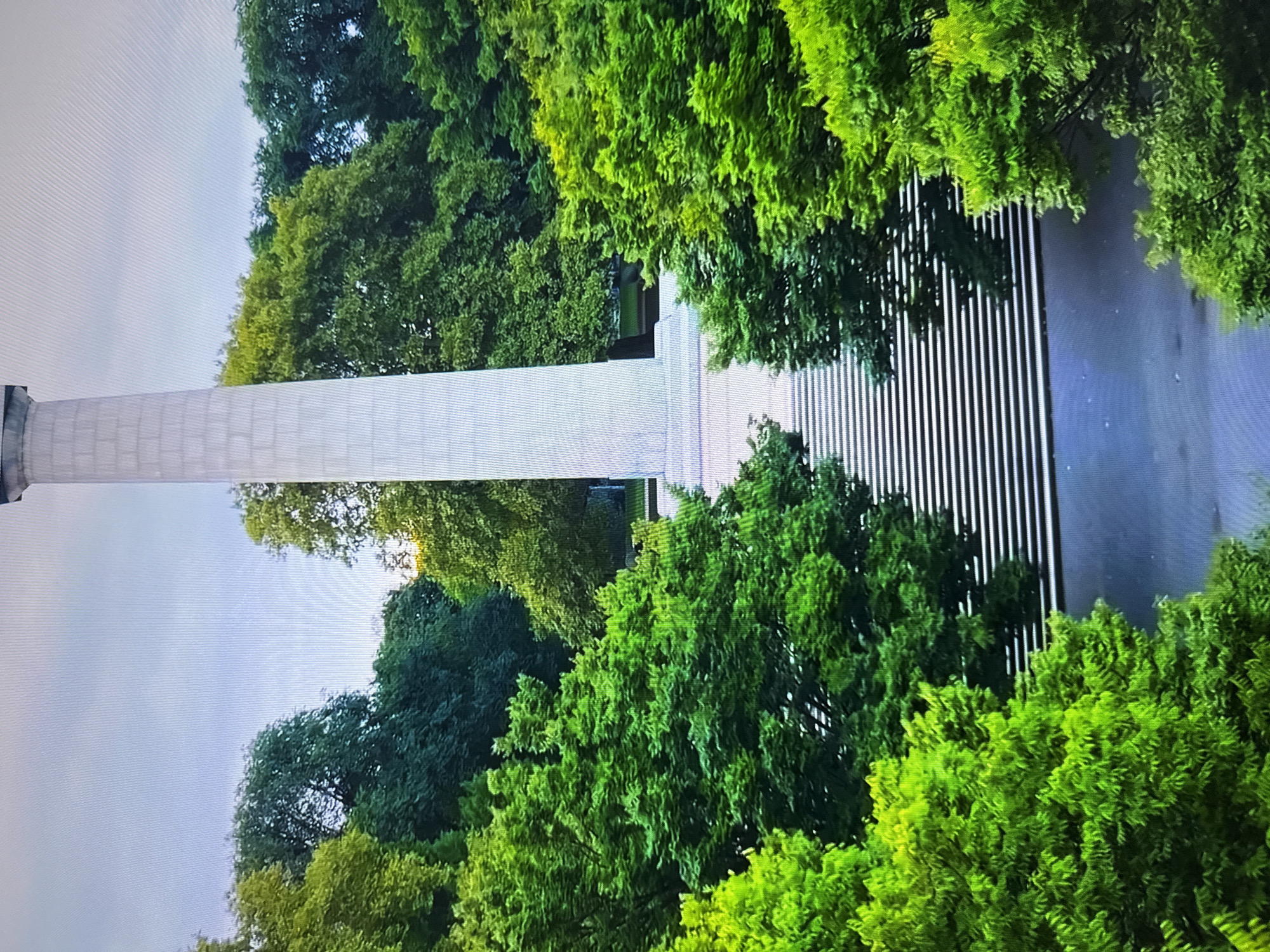 The image shows a tall, light-colored monument, likely a column or obelisk, surrounded by lush green trees. A wide staircase with white steps leads up to the base of the monument. The sky above is overcast and pale. The scene appears to be in a park or public square within the city of Burgas, Bulgaria, as suggested by the monument's style and the surrounding foliage which is typical of European city parks. There are no people visible in the frame.