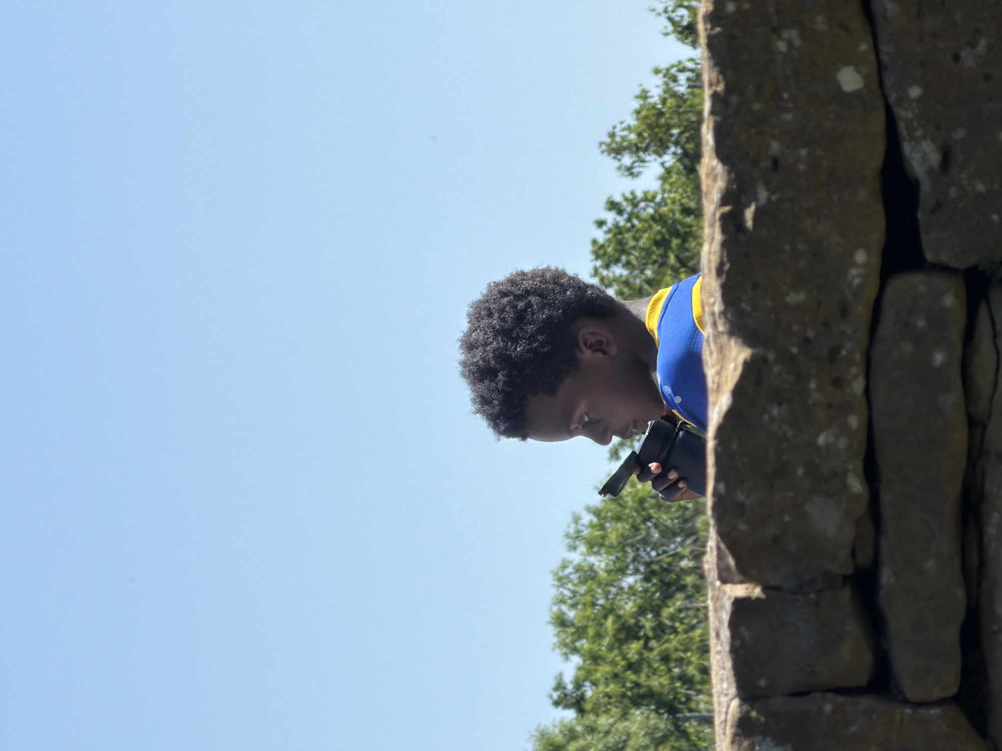 A low-angle, medium shot shows a dark-skinned young man peering into a black instrument. He is positioned behind a rough-textured stone wall that angles vertically. The man has short, dark curly hair, and he is wearing a blue shirt with a yellow shoulder stripe. The backdrop includes a clear blue sky and the foliage of green trees. The location is the city of Fentange, Luxembourg.