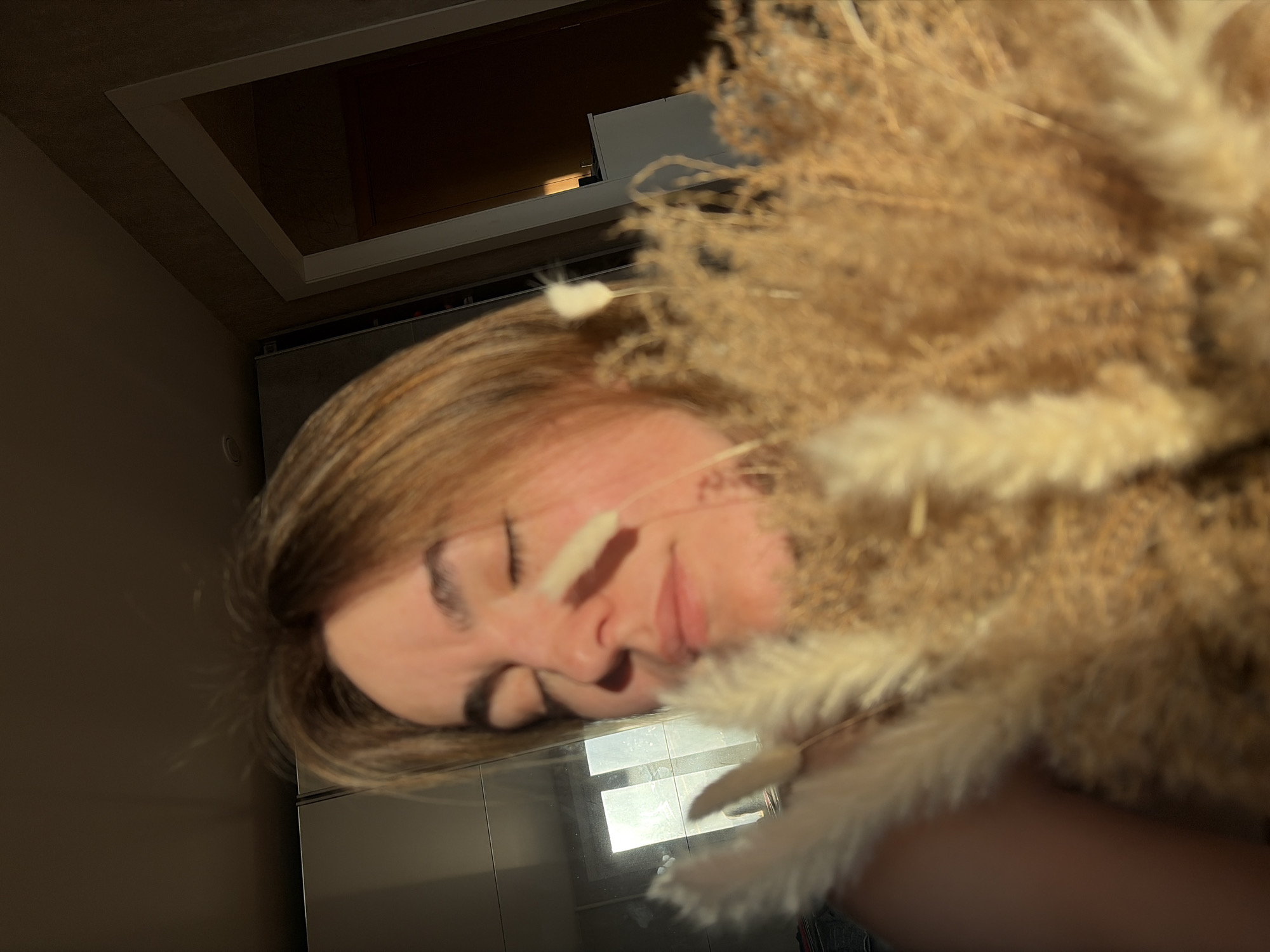 The image shows a woman holding a bouquet of dried flowers in an interior space. The location is likely a room in a building in Izhevsk, Russia. The woman has light brown hair and closed eyes. Her face is partially obscured by the bouquet. Behind her, there is a wall and what appears to be a reflective surface with a window. The lighting is bright, possibly from sunlight. There is also a ceiling with a recessed light fixture.