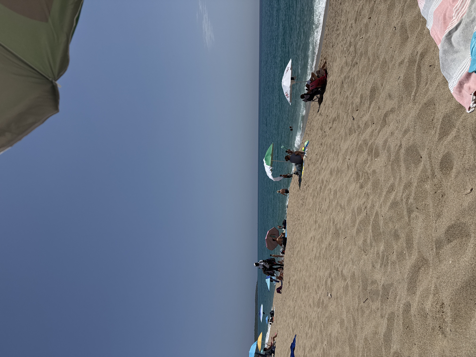 The image depicts a crowded beach scene on a sunny day. The foreground is dominated by sand, with some beach towels and debris scattered on it. In the middle ground, several beach umbrellas provide shade for sunbathers. A person is seen standing near some umbrellas, possibly a vendor. People are scattered across the sand, some sitting, some standing, and some are in the shallow water. The sea is a vibrant blue with gentle waves. The horizon is visible, with a clear blue sky above. A small island or peninsula is visible in the distance on the left. The scene suggests a typical summer day at a popular beach resort.