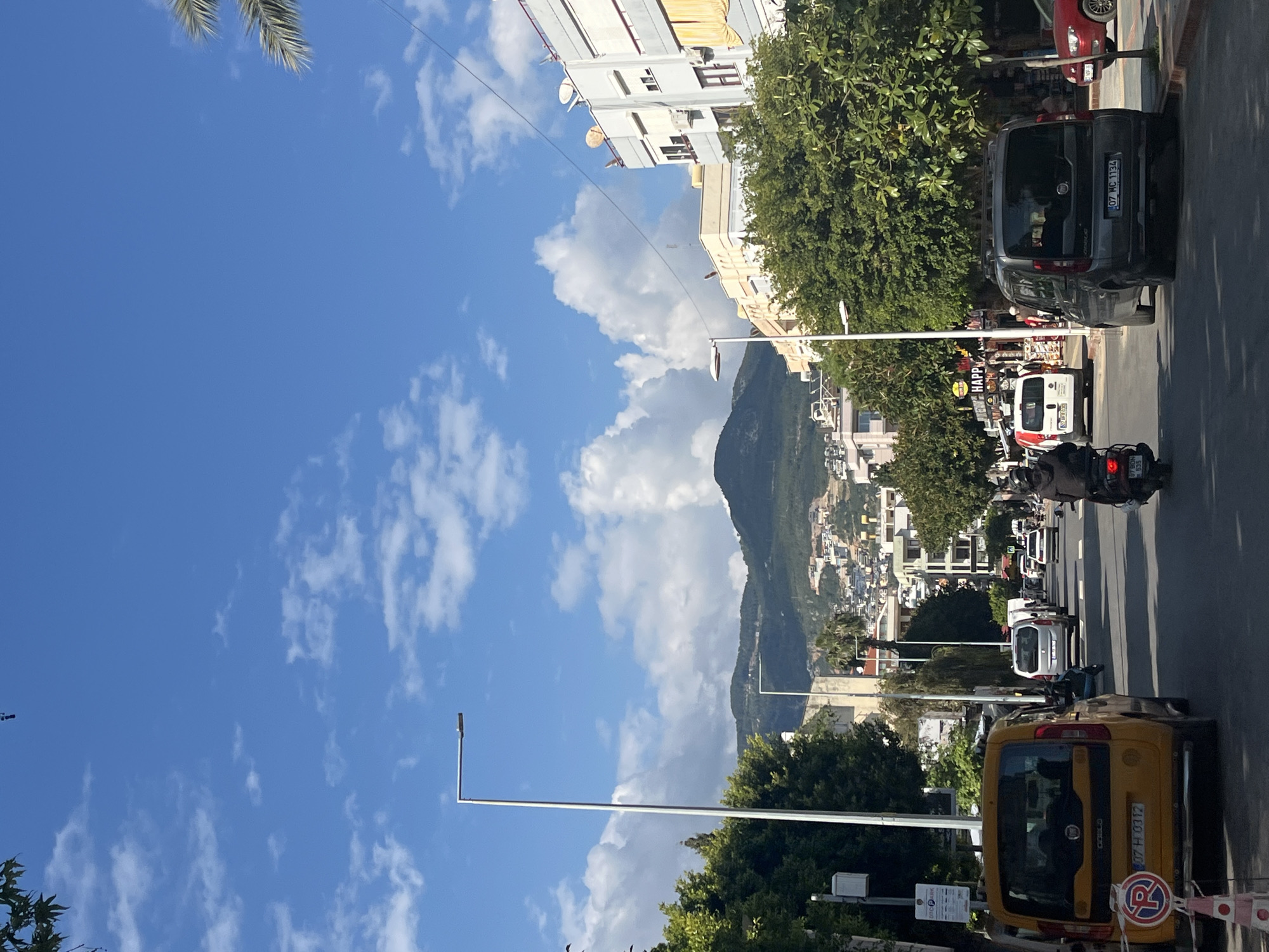 The image depicts a street scene in Alanya, Turkey. The view is looking uphill towards a mountain partially obscured by clouds. The sky is blue with scattered white clouds. Along the right side of the road, there is a line of vehicles, including cars, vans, and a motorcycle. Some parked and others moving. Trees line the roadside, and buildings are visible in the background. A yellow taxi is in the foreground. Streetlights are also visible. The general environment suggests a sunny day in a populated area of Alanya.