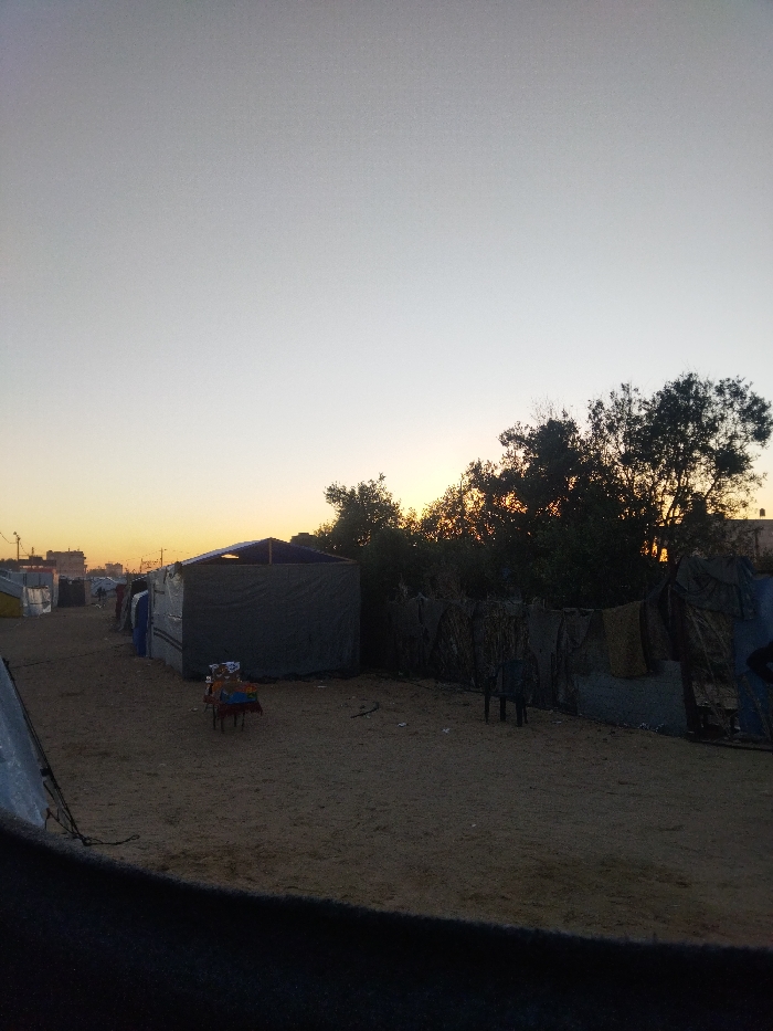 The image depicts a desolate outdoor scene in Khan Yunis, Palestinian Territory, during either sunrise or sunset, evidenced by the gradient sky transitioning from light blue-grey overhead to an orange-yellow glow at the horizon. The ground consists of uneven light brown sand, sparsely scattered with small debris. On the left, a line of temporary shelters or tents extends into the distance, constructed from tarps and various materials; the closest is a dark grey structure with a peaked roof. Further along this line, other shelters are visible, some with reflective surfaces, and distant urban structures are faintly silhouetted against the horizon. In the mid-ground, a low, red-framed table holds an assortment of colorful items. To the right, a dense thicket of trees and brushwood stands in silhouette, largely obscuring the direct source of light. In front of the trees, a makeshift barrier or fence is erected from fabric, tarpaulins, and interwoven brushwood, with several pieces of fabric or clothing hanging on it. A single dark plastic chair rests on the sand in this area. The immediate foreground is partially obscured by a dark, indeterminate object, possibly the edge of another shelter or fabric. No individuals are present in the frame.