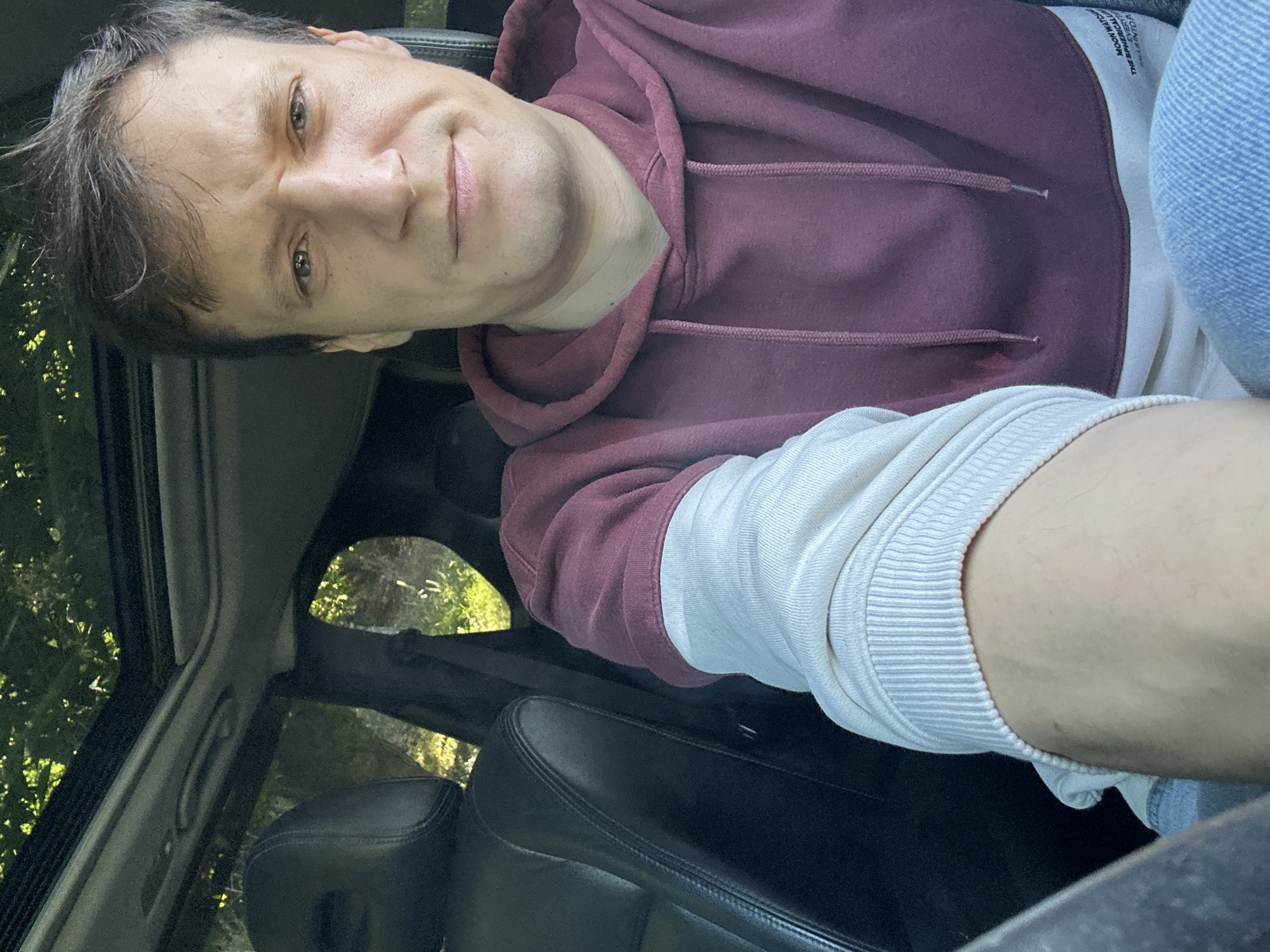 A man is taking a selfie inside a car. He is wearing a burgundy and white hooded sweatshirt and blue jeans. The car has a sunroof and the interior is visible, along with the exterior surroundings of trees and foliage, suggesting a rural or park-like setting. The man's facial expression is neutral, and he is looking directly at the camera. The image does not provide any specific indicators of the city of Makiv, Ukraine.