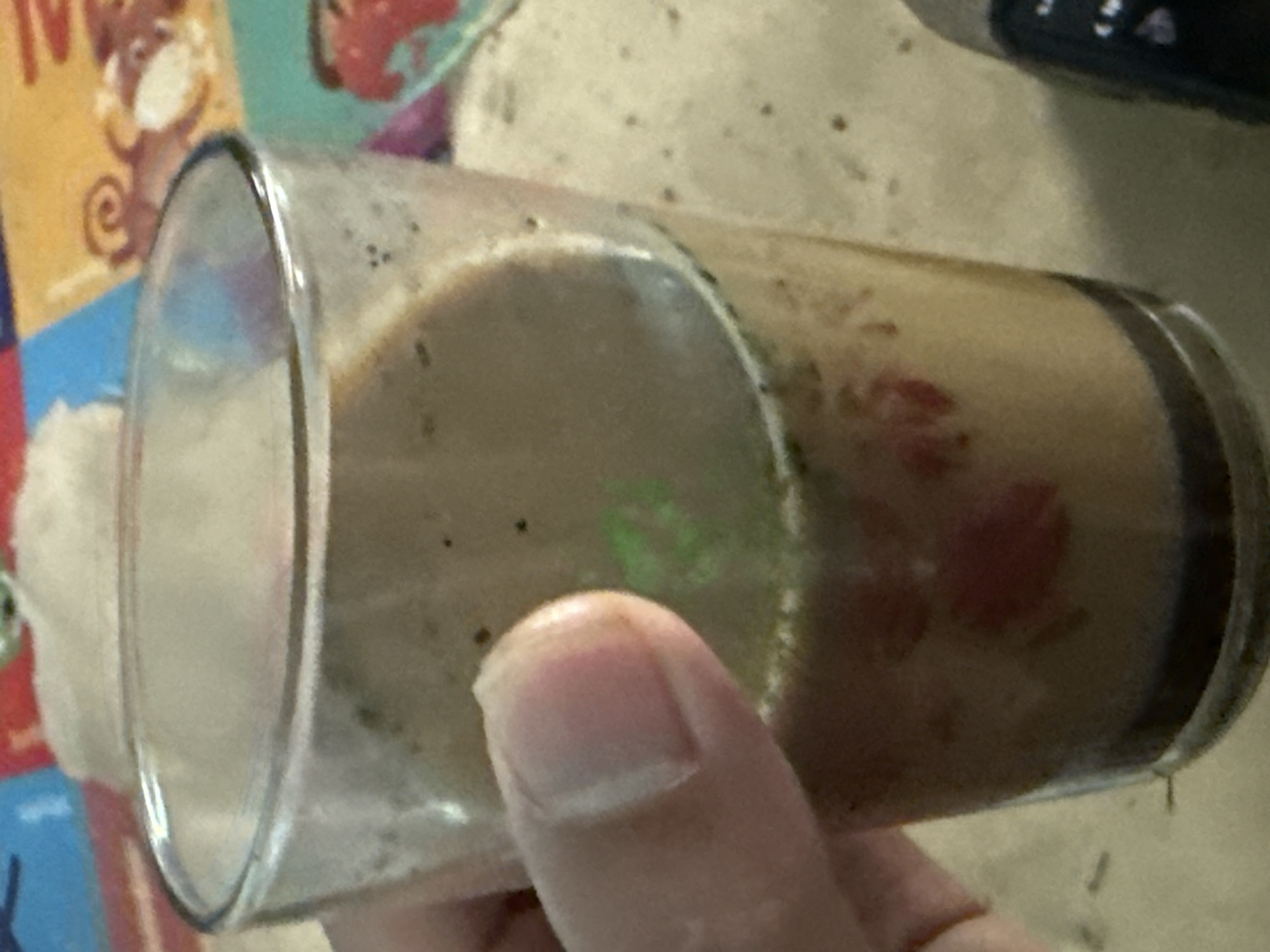 A glass of a brownish beverage with small dark specks is held by a hand with a visible thumb. The beverage appears to have some red pieces and a faint green swirl. The background shows a blurry surface with patterns, possibly a tablecloth or placemat, with some scattered dark specks. A portion of a black electronic device is visible on the right side of the image. The image is a close-up shot, focusing on the glass and the hand. There is no discernible indication of the city of Slahung, Indonesia, in this image.