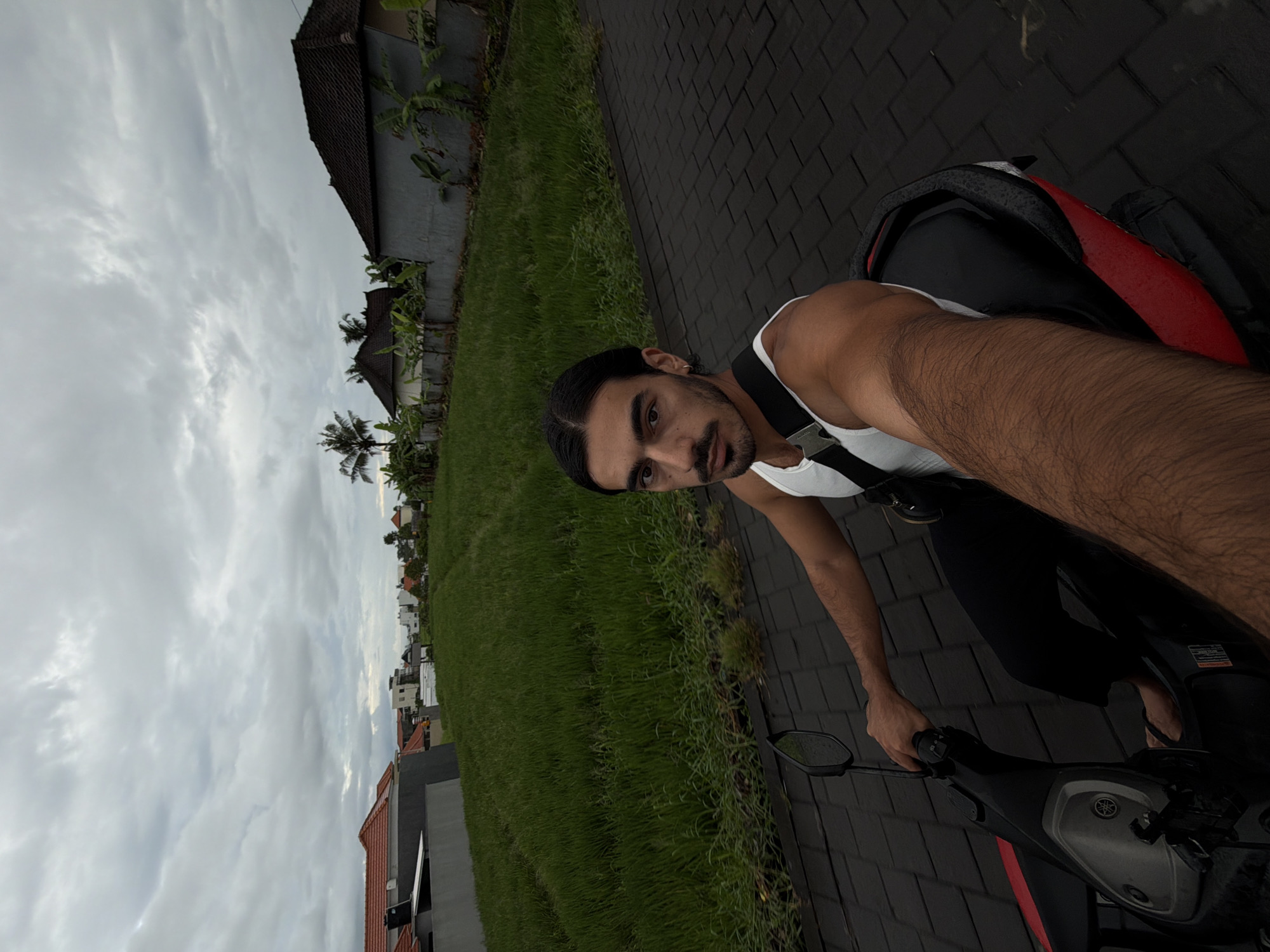 A man is captured in a selfie-style shot while seated on a scooter on a cloudy day in what appears to be a rural or semi-rural area of North Kuta, Indonesia. The man, with dark hair pulled back and a beard, is wearing a white tank top and dark pants, and looks directly at the camera with a neutral expression. His right arm is visible, holding the scooter's handlebar, while his left arm is not fully in view. A red and black backpack rests on the scooter's seat behind him.
The scooter itself is dark-colored with red accents, and the Yamaha logo is visible on its front panel. The paved path beneath the scooter is made of dark interlocking bricks. To the left of the path stretches a vibrant green field, likely a paddy field, with scattered houses and palm trees in the background. More buildings with traditional-style roofs are visible along the path and in the distance. The sky is overcast with light gray clouds, indicating a cloudy day, possibly morning or late afternoon, as there are no distinct shadows. The overall scene suggests a leisurely moment amidst the natural and architectural landscape typical of the specified region.