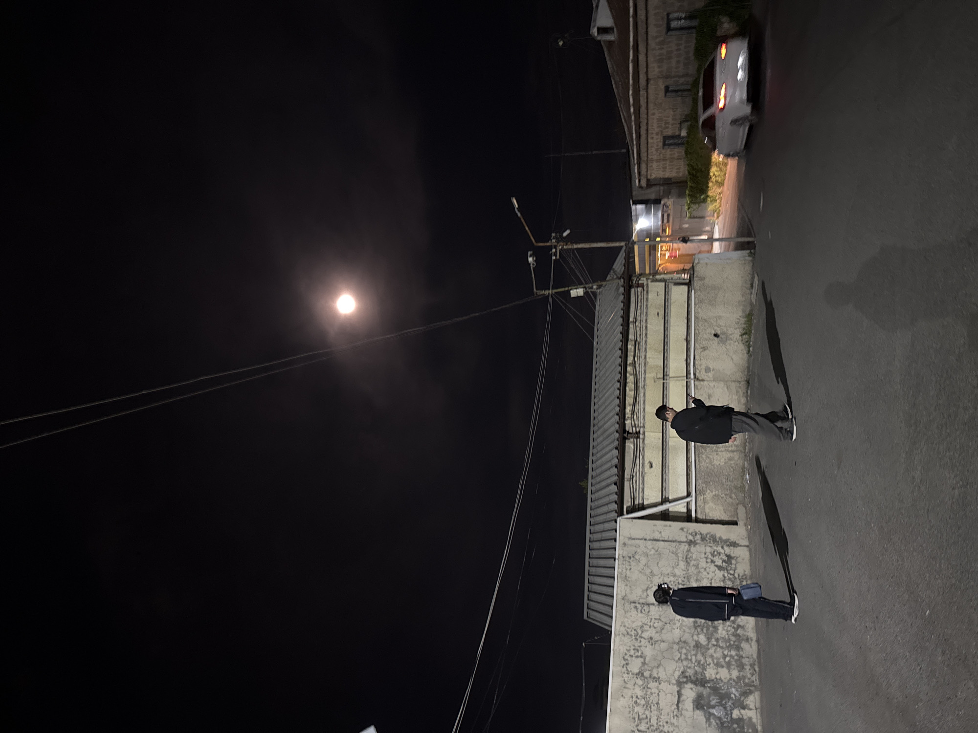The image depicts an urban or semi-urban street scene at night, set in Yerevan, Armenia. The photograph is oriented vertically, with the ground extending from the bottom to the top right of the frame, and the sky occupying the left side.
Two individuals are standing on a paved street or alleyway. The person closer to the camera is seen from behind, wearing a dark jacket, dark pants, and light-colored shoes, with a light blue shoulder bag. They cast a long shadow stretching to their left. Further down the street, a second person is also standing, facing away from the camera, dressed in a similar dark attire with light shoes, and also casting a long shadow.
On the right side of the street are various buildings, featuring stone or brickwork and concrete walls, with some corrugated metal siding visible. Further down the street, a white car is partially visible, appearing somewhat blurred, suggesting motion or a slight camera shake.
The sky is dark, indicating nighttime, but a very bright, circular light source, likely the moon, is prominent in the upper left portion of the sky, illuminating the scene and creating the long shadows. Overhead utility wires traverse the sky, connected to a utility pole structure on the right, which also has what appear to be streetlights mounted. The overall atmosphere is dark, with artificial light sources providing limited illumination beyond the primary bright sky object. There is no visible text in the image.
