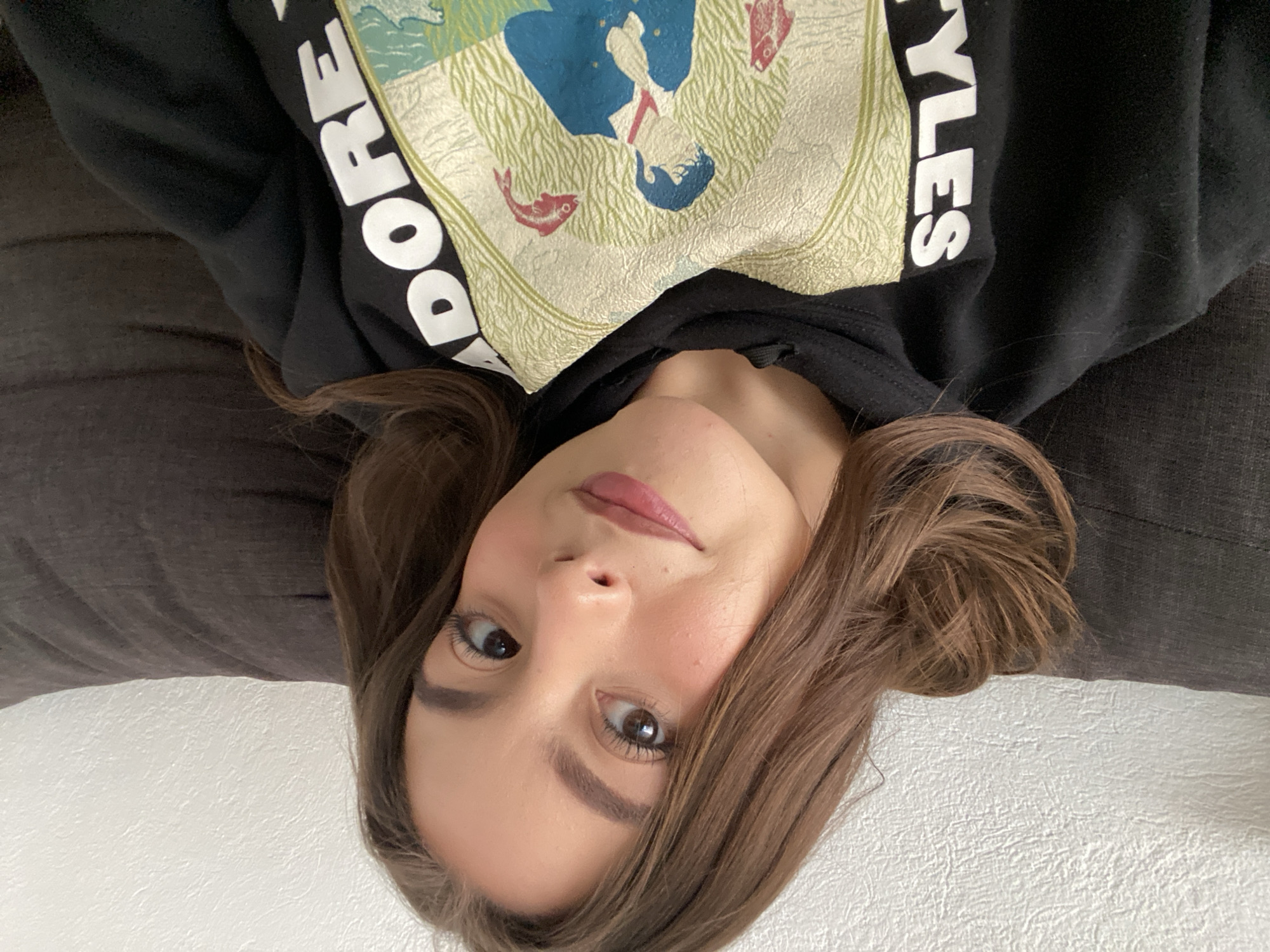 A person is pictured in an upside-down perspective, lying down on a dark gray fabric surface, possibly a couch or bedding. They are wearing a black hooded sweatshirt with white lettering reading "DORE STYLES" on the sleeve. The front of the sweatshirt features a graphic design with blue, green, and red elements. The person has brown hair styled with bangs, light skin, and is wearing subtle makeup. A white textured surface is visible in the background. The location is unknown.