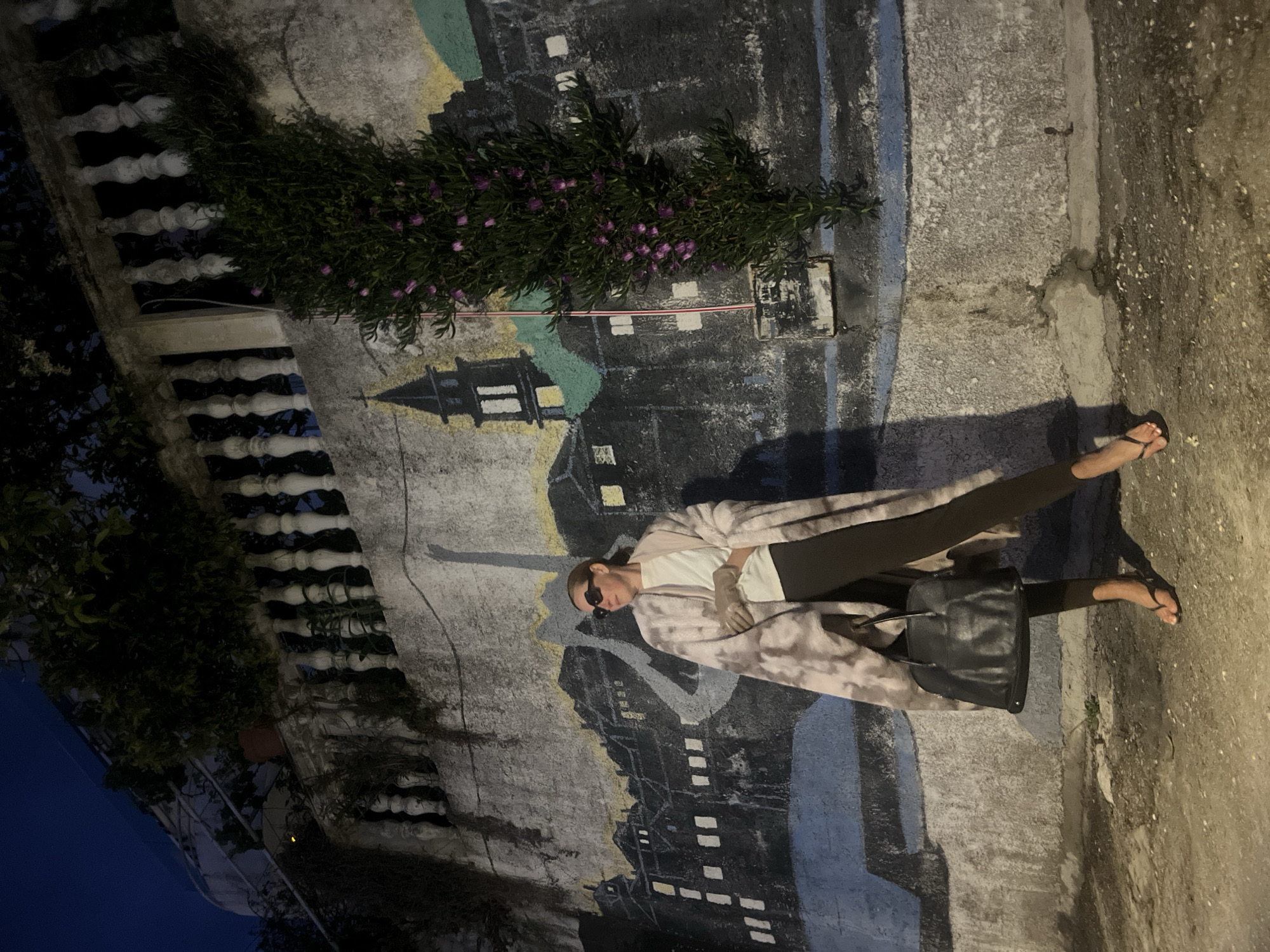 A nighttime photograph, rotated 90 degrees clockwise, captures a person standing casually on a concrete path or sidewalk in Budva, Montenegro, beside a painted wall mural.
The subject is a female figure, centered in the frame, wearing dark sunglasses, a white t-shirt, dark leggings, and a long, light-colored, possibly tie-dye patterned cardigan or coat. She has her hair tied back and is wearing black flip-flops. A black shoulder bag hangs from her left arm. She stands relaxed, with one foot slightly ahead of the other, posing for the camera. A long shadow of the person is cast on the ground to her right, indicating an off-camera light source.
Behind the person is a large mural on a textured concrete wall, depicting a stylized cityscape or landscape. The mural features dark buildings with illuminated white windows, a prominent tower (resembling a pagoda or ornate steeple), and sections of light yellow/beige, black, and blue paint. Above the mural, a dense green bush with small purple flowers extends along the wall. To the left of the bush, a white, ornate concrete balustrade is visible, suggesting a balcony or an elevated structure. The sky visible through the balustrade is dark blue, confirming it is nighttime. The ground appears to be made of concrete or asphalt.
There is no visible text in the image. The overall impression is a casual nighttime portrait in an urban setting.