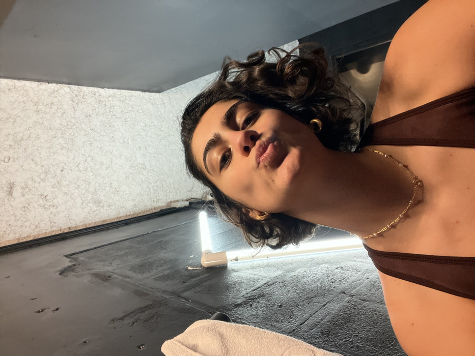 A woman wearing a brown halter top and gold jewelry poses from a low angle. She has short, dark curly hair, tan skin, and is puckering her lips toward the camera. Behind her is a glossy black floor and a textured white wall. A long, thin white light is visible in the background, illuminating part of the floor and wall. A white towel is partially visible at the bottom of the frame. The lighting creates shadows and highlights across the scene.