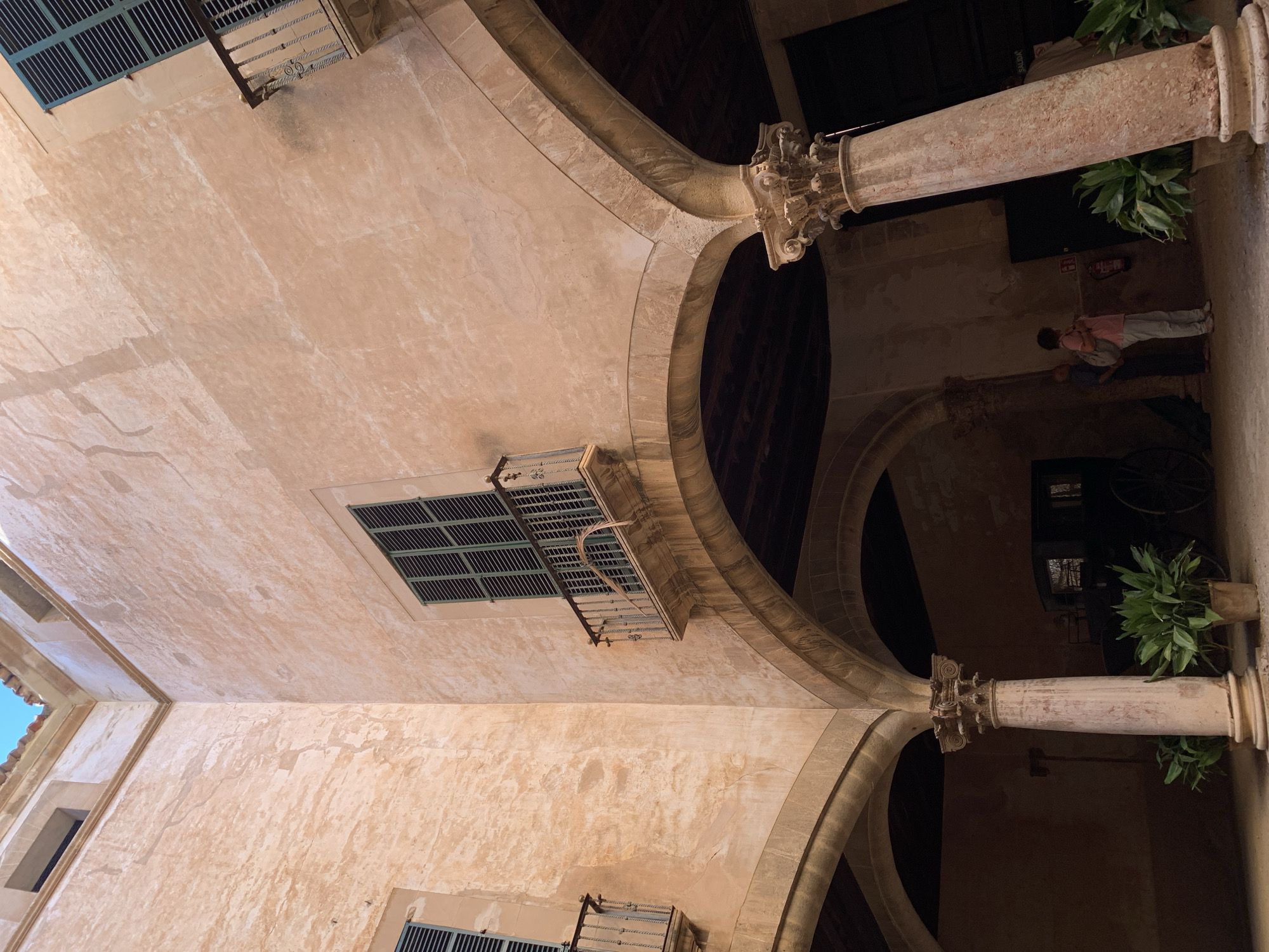 The image depicts an interior courtyard of a building in Palma de Mallorca, Spain. The architecture features warm, peach-colored stucco walls, arched walkways supported by classical columns with ornate capitals, and wooden beamed ceilings. Several windows with green shutters and wrought-iron balconies are visible on the upper floors.
In the lower part of the courtyard, two potted green plants flank a large, light-colored column. Behind the column, a dark-colored horse-drawn carriage is partially visible. Two individuals are standing near the carriage, facing away from the camera. One person is wearing a pink shirt and light-colored pants, carrying a backpack, and looking upwards. The other person is wearing a blue shirt and dark pants, also looking upwards. A fire extinguisher is mounted on the wall to the left of these individuals. A sign that reads "SALIDA" (Spanish for "Exit") is visible on the wall in the background. The lighting suggests it is daytime, with sunlight illuminating parts of the courtyard and casting shadows. The overall impression is one of historical architecture and a tranquil, shaded interior space.