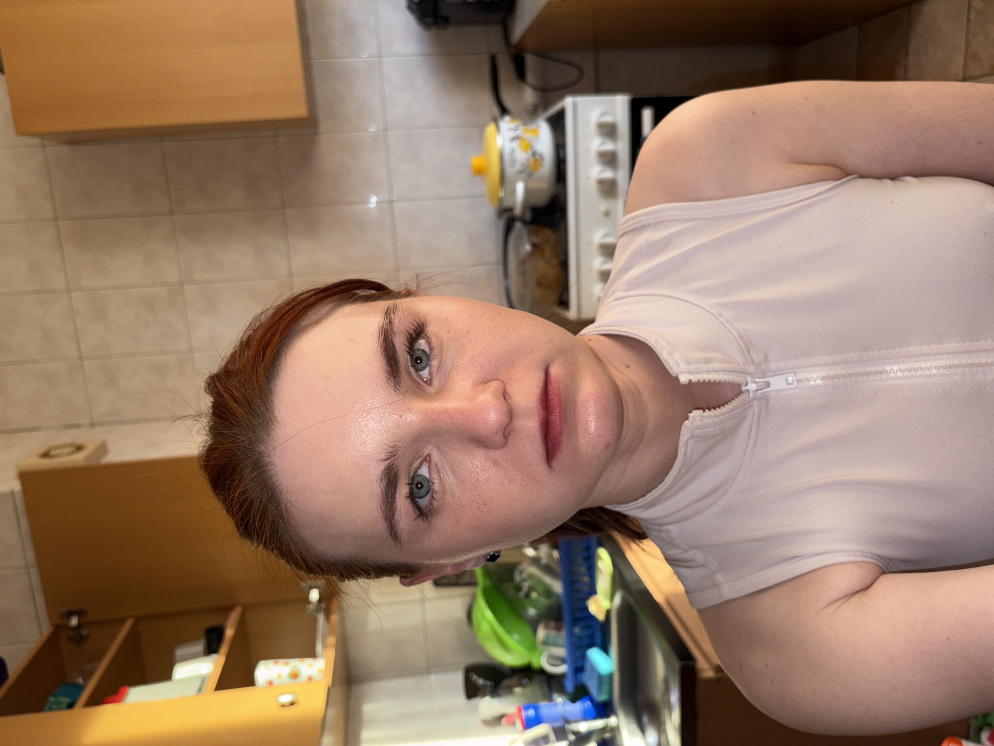 A close-up, rotated portrait shows an individual, likely female, in what appears to be a kitchen in Unknown, Russia. The person has reddish-brown hair and blue eyes, and their face exhibits noticeable swelling, particularly around the lips and chin. Their expression appears serious or possibly distressed, and the skin has a somewhat glossy appearance.
The subject is wearing a light-colored, possibly off-white or light pink, short-sleeved top with a prominent white zipper down the front.
The background details a kitchen environment with light-colored square tiles on the wall. Light brown wooden cabinets are visible both above and below. To the upper right, a white stove or oven with control knobs is partially in view, along with a small yellow-lidded pot decorated with a floral pattern. Further to the right, a sink area, a green bowl, and a blue dish rack can be discerned. Another wooden cabinet, slightly ajar and containing various household items, is visible in the lower left. The scene is illuminated by indoor artificial lighting. No visible text is present in the image.