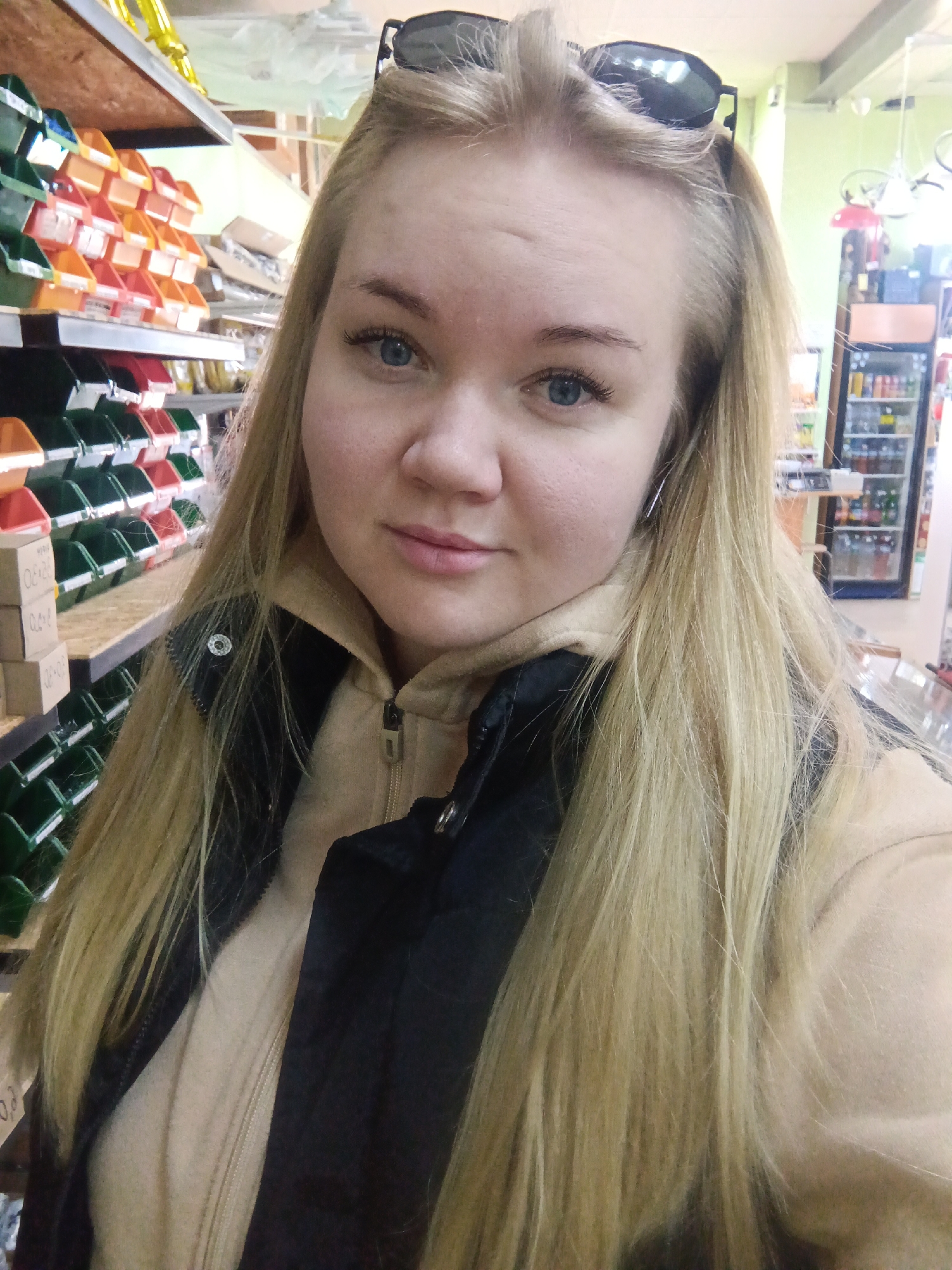 A young woman with long blonde hair and blue eyes is taking a selfie indoors. She has sunglasses on her head and is wearing a tan hoodie underneath a black vest. She is standing near a shelf filled with colorful plastic bins and cardboard boxes. In the background, there is a beverage cooler with drinks visible inside. The location is Фоки, Russia.