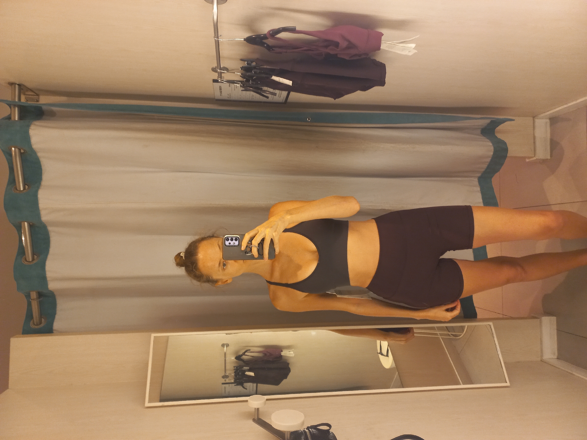 A woman is standing in a changing room, taking a photo of herself in the mirror with her smartphone. She is wearing a dark-colored athletic top and shorts. Above her, several articles of clothing hang on a rack attached to the wall. A mirror is positioned beneath the curtain. The setting appears to be a retail clothing store or department. There are no specific indicators to confirm the location is in Pattaya City, Thailand.
