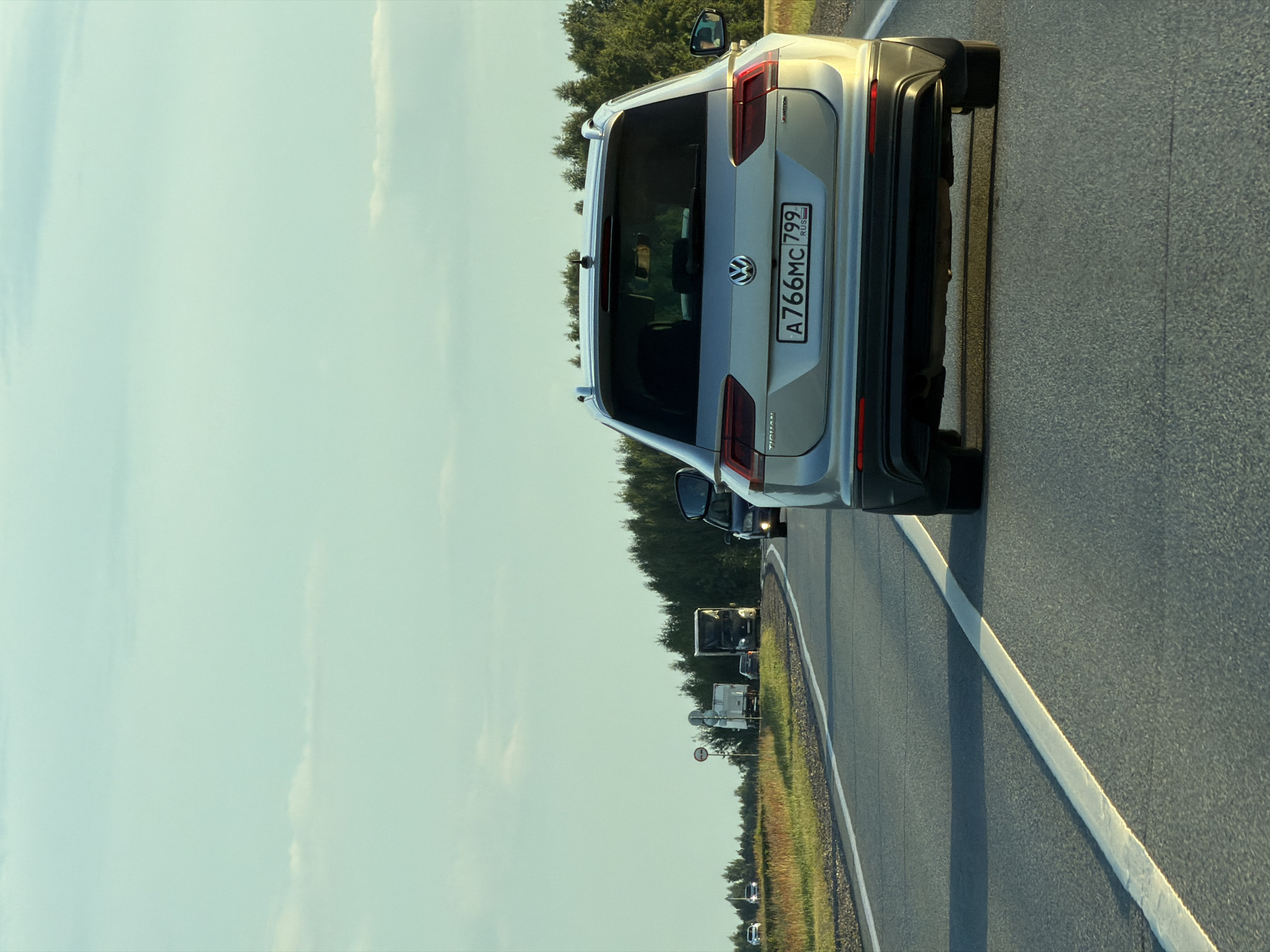 A silver Volkswagen Tiguan, bearing a Russian license plate A766MC799, is seen from the rear driving on a two-lane asphalt road. The car is in the right lane, followed by other vehicles. The road is bordered by a grassy shoulder and a dense forest of green trees on the right. In the distance, there are other vehicles including trucks and cars, and a road sign is visible on the left side of the road. The sky is a light blue with sparse clouds, indicating daylight. The scene appears to be a rural or suburban highway, possibly in or near the city of Obukhovo, Russia, given the license plate and surrounding landscape. The lighting suggests it is late afternoon or early evening.