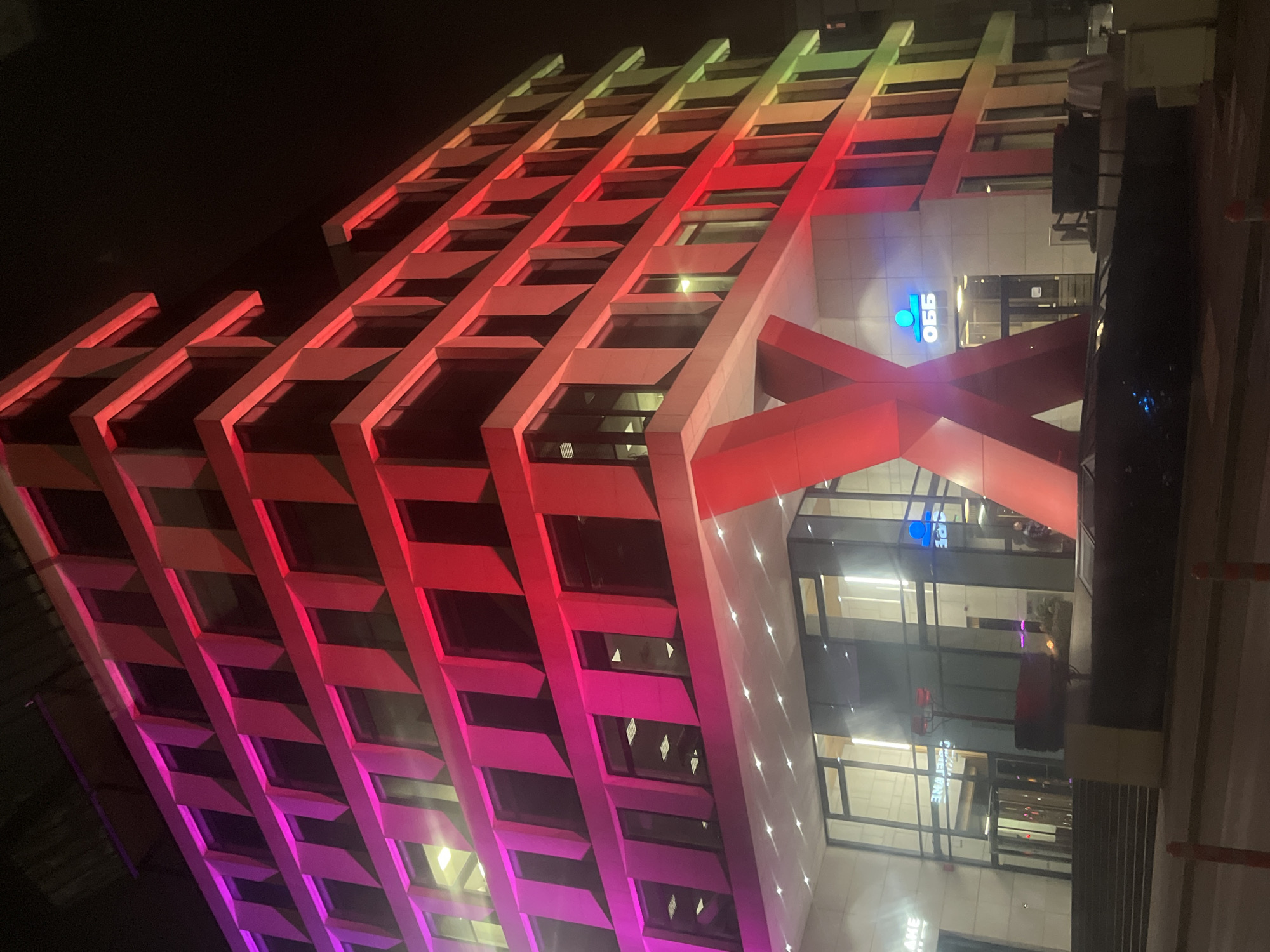 A nighttime view of the OBB (UniCredit Bulbank) building in Sofia, Bulgaria, showcases its architectural features and unique lighting. The building's facade consists of a multi-story structure with horizontal and vertical elements, forming rectangular sections. These elements are illuminated with a gradient of colors, transitioning from green and yellow at the top to red and purple toward the bottom. At the base of the building, a prominent "X"-shaped support is lit in red. The building's entrance area, marked by the OBB logo, features large glass windows that reflect the interior lighting. The surrounding environment appears to be urban, with dark skies and ambient lighting from nearby structures.