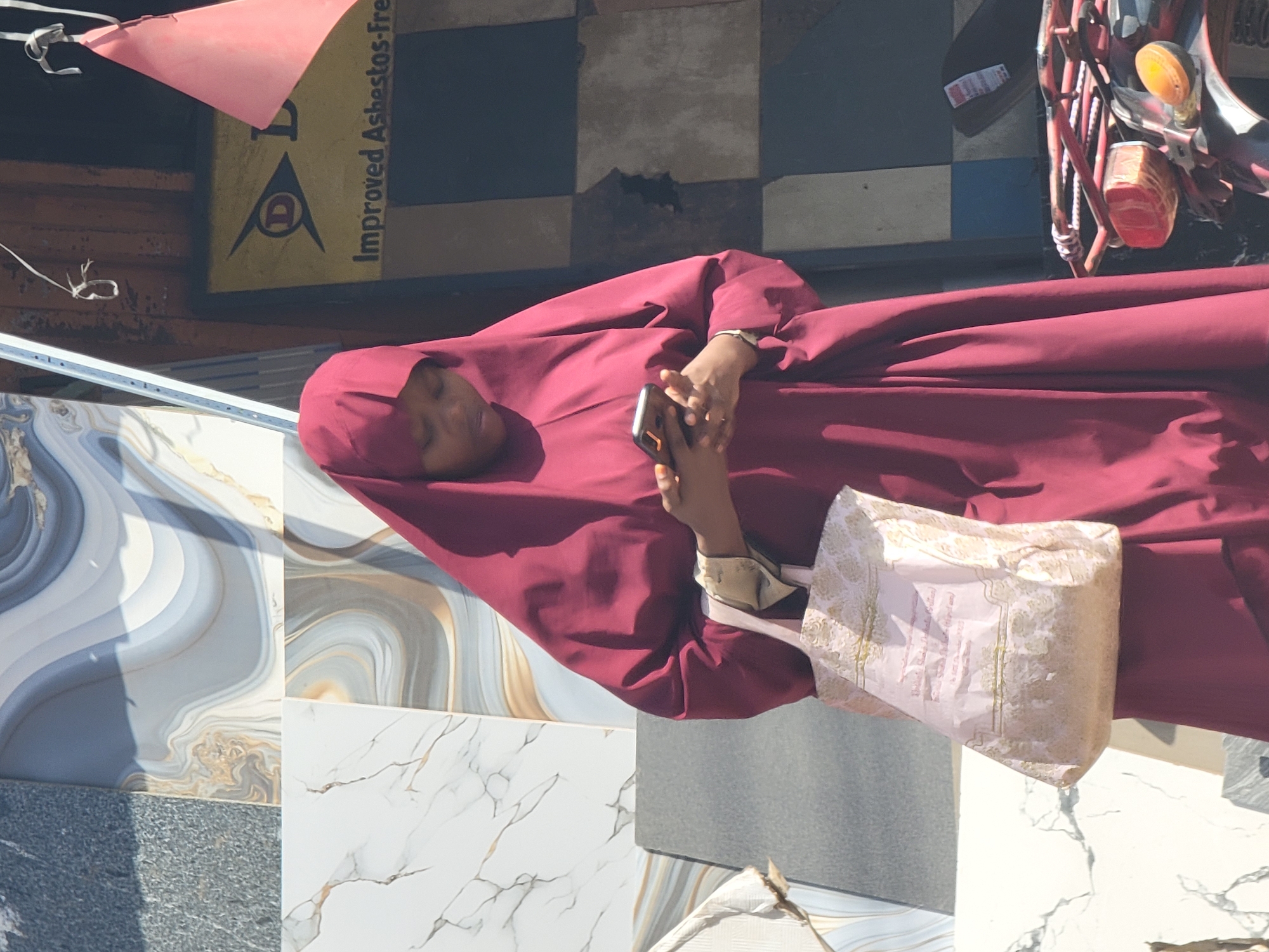 A woman wearing a maroon hijab is holding a smartphone in her hands and is standing in front of a display of various floor tiles in Maiburiji, Nigeria. She is also holding a white gift bag with gold trim. Behind her, there's a moped parked in front of a storefront with a sign that reads "Improved Asbestos-Free." The environment appears to be outdoors during daylight.