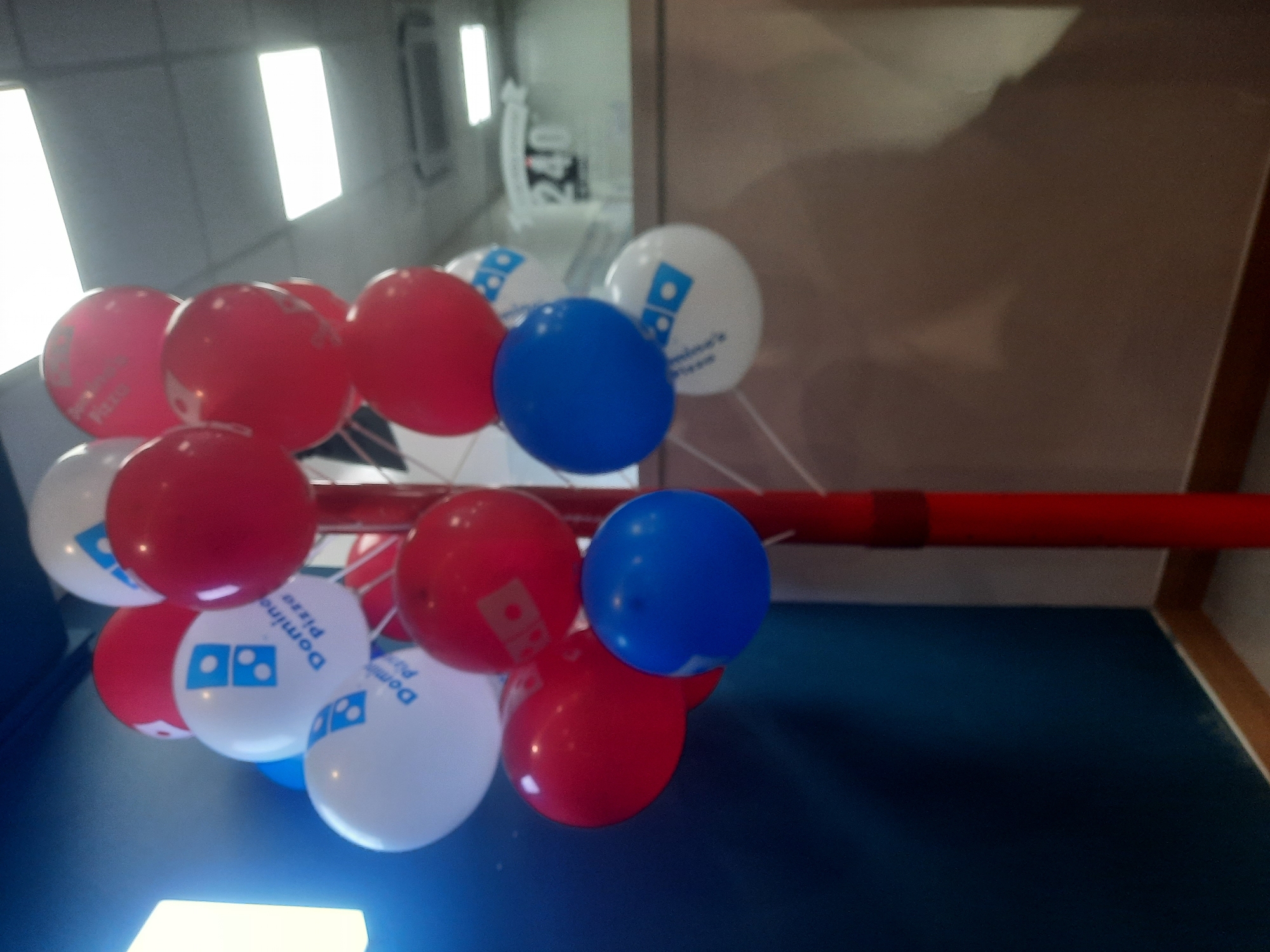 The image shows a cluster of Domino's Pizza branded balloons, predominantly red, white, and blue, attached to a red pole. Some of the white balloons feature the Domino's Pizza logo. The balloons are arranged in a cascade. The background suggests an indoor commercial setting, with a dark blue wall on the left, and a blurred background with what appears to be a ceiling with fluorescent lights and a sign with the number "240" visible in the distance. There are no people visible in the image. The lighting suggests it is daytime or the interior lights are on. The overall scene depicts a festive or promotional display for Domino's Pizza, potentially in a store or at an event. The specific location in North Jakarta, Indonesia, cannot be determined from the visual information alone.
