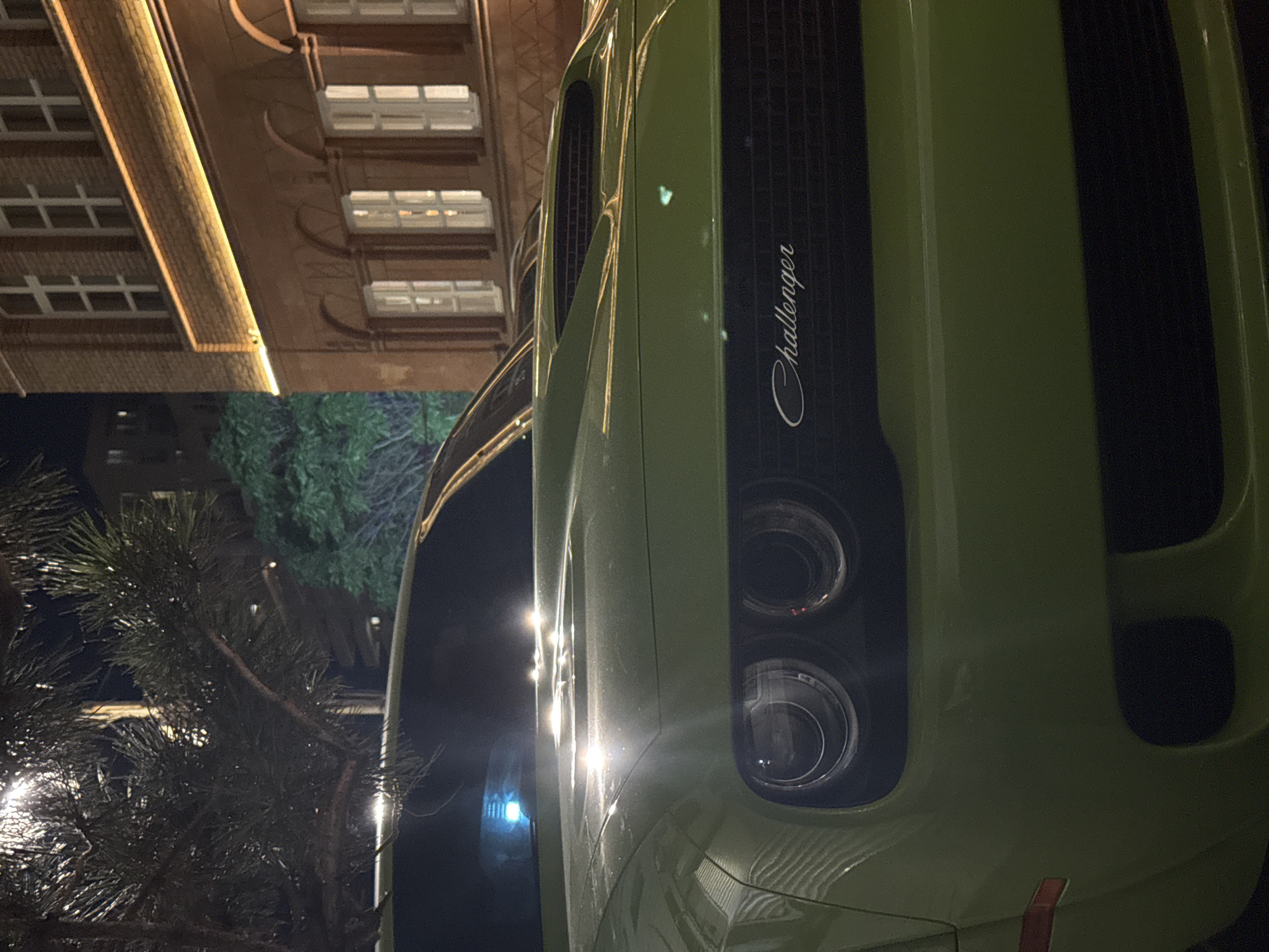 The image shows the front of a lime green Dodge Challenger parked at night in a city. The car's headlights and hood are visible, as is the "Challenger" emblem on the grille. In the background, a brick building with arched windows and exterior lighting is illuminated. Trees with dark foliage are also present. The scene appears to be a street or parking area in Yerevan, Armenia, given the architectural style of the building and the general urban ambiance. There are no people visible in the image.