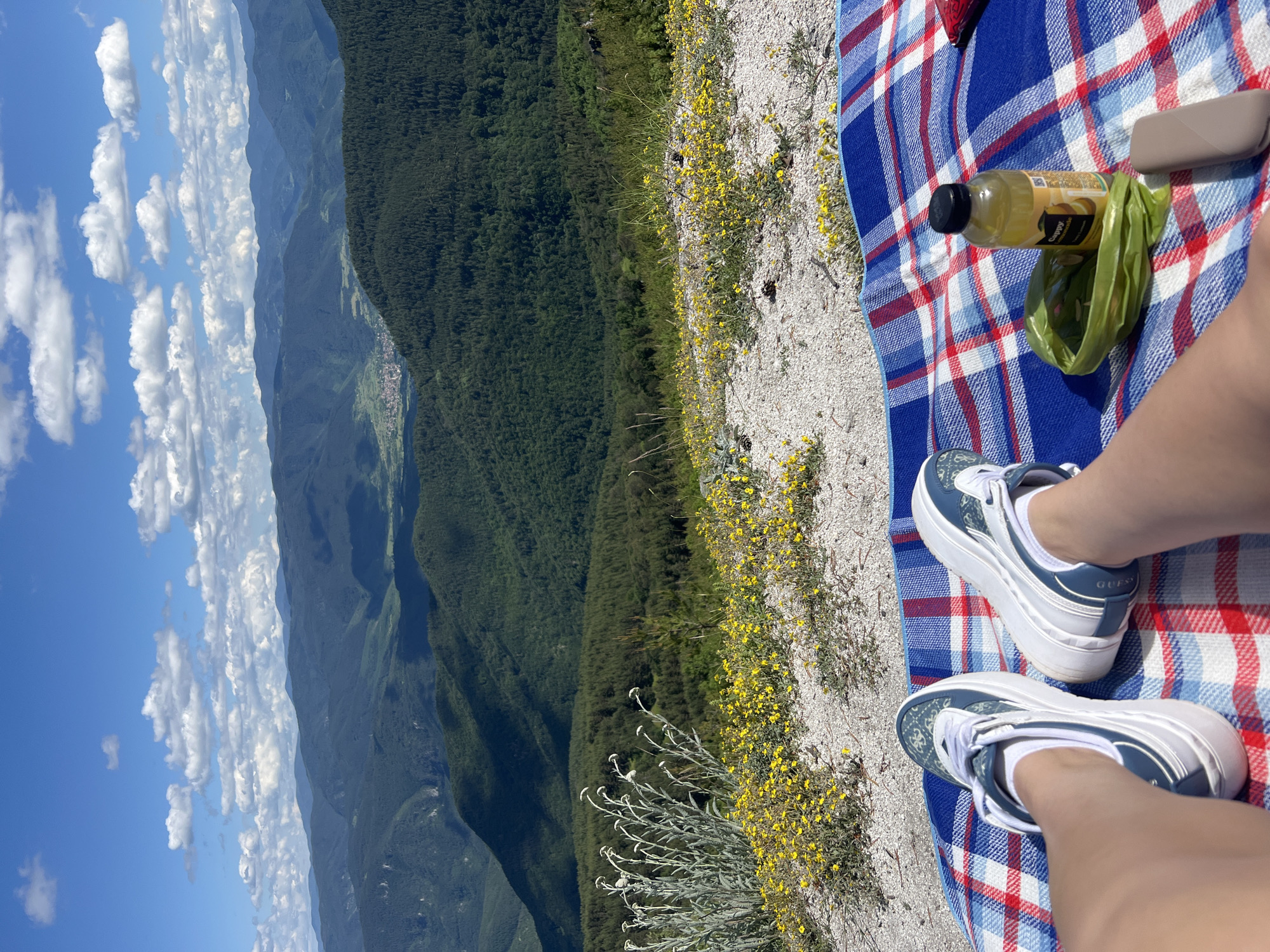 The image depicts a person's legs and feet wearing white and blue sneakers, resting on a red, white, and blue checkered blanket. To the right of the legs are a bottle of yellow liquid and a green bag. The blanket is laid out on a rocky, sandy surface covered with small yellow flowers. In the background, a lush green mountainous landscape unfolds under a bright blue sky dotted with white clouds. The location is identified as Kosovo, Bulgaria, suggesting an outdoor recreational setting.
