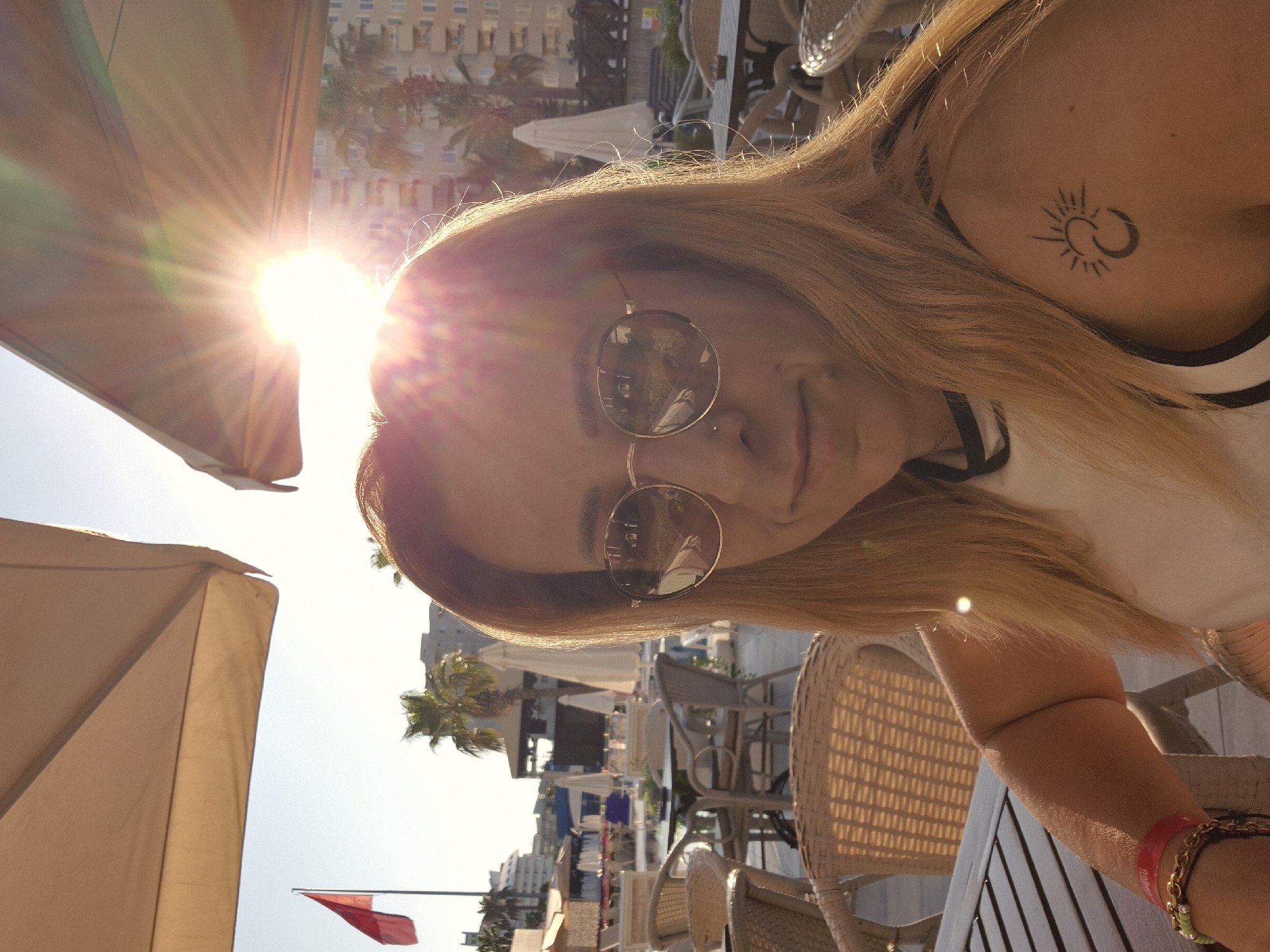 In a selfie taken outdoors, a woman with blonde hair and sunglasses smiles at the camera. She has a nose ring and a tattoo of a sun and moon on her left shoulder. She is wearing a white top with black trim and a red bracelet on her left wrist. In the background, there are palm trees, buildings, and outdoor seating areas with tables and chairs. The sun is shining brightly, creating a lens flare effect.