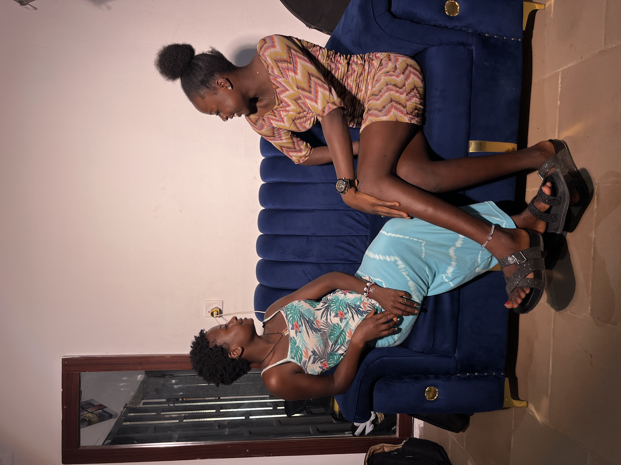 Two women are seated on a plush, dark blue couch in an indoor setting. The woman on the left is wearing a sleeveless dress with a tropical floral print and a light blue tie-dye skirt. She has short, curly hair and is looking to her right. She is adorned with several bracelets and a ring. The woman on the right is wearing a short, patterned dress with a chevron design and her hair is pulled up into a bun. She is looking to her left and is wearing a watch on her left wrist. They are positioned facing each other, suggesting they are engaged in conversation. The couch has gold accents on its legs and decorative trim. The background shows a wall with an electrical outlet and a framed mirror. The floor appears to be tiled. The lighting is dim, creating shadows.
