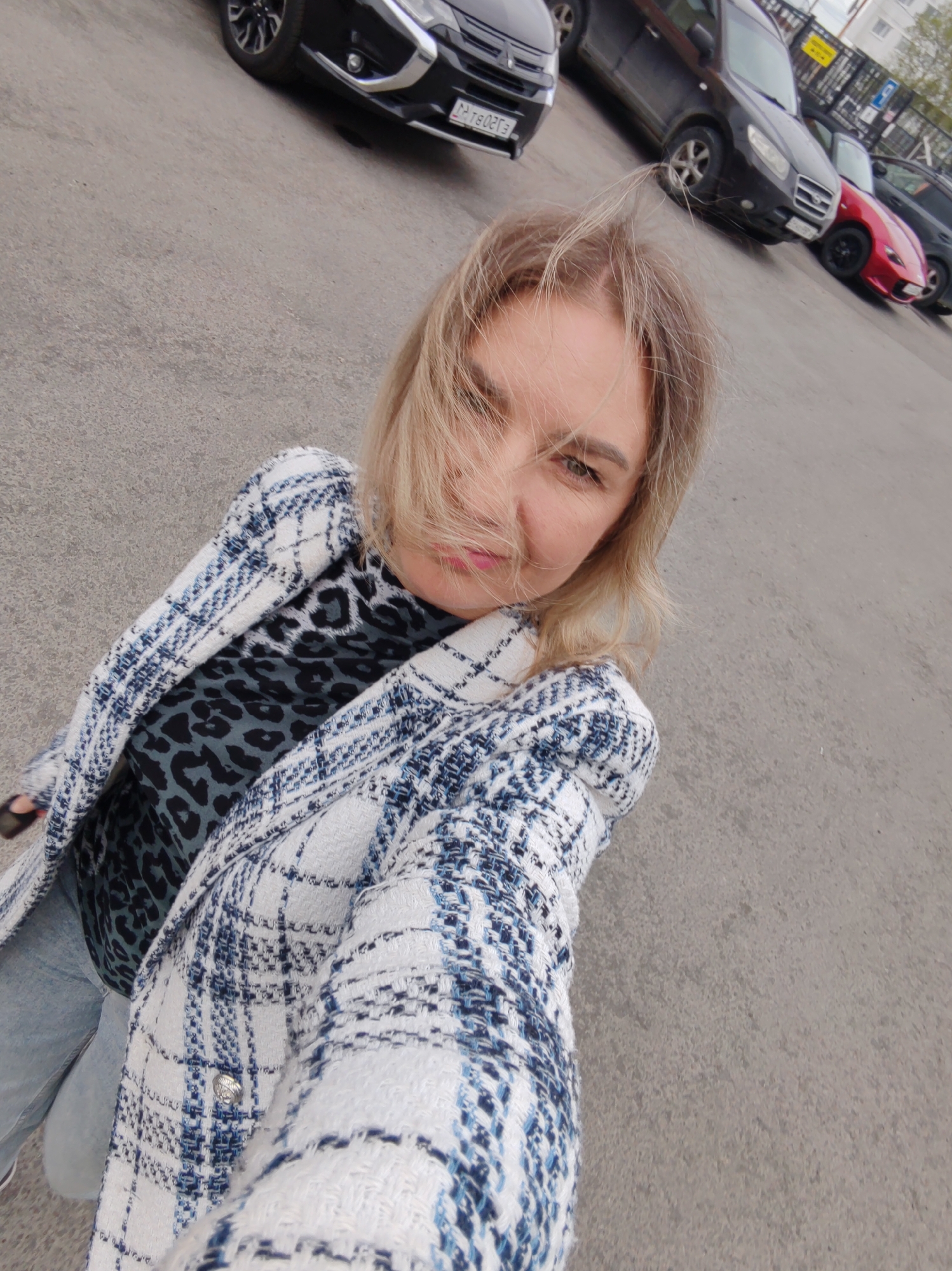 A blonde woman is standing outside in a parking lot. She is wearing a white and blue checkered jacket over a leopard print shirt, and jeans. Her hair is blowing in the wind, partially obscuring her face. In the background, several cars are parked, including a black SUV and a red sports car. The scene appears to be in Petropavlovsk-Kamchatsky, Russia, suggested by the plate number on the black SUV that reads "PTB027E". The ground is asphalt.