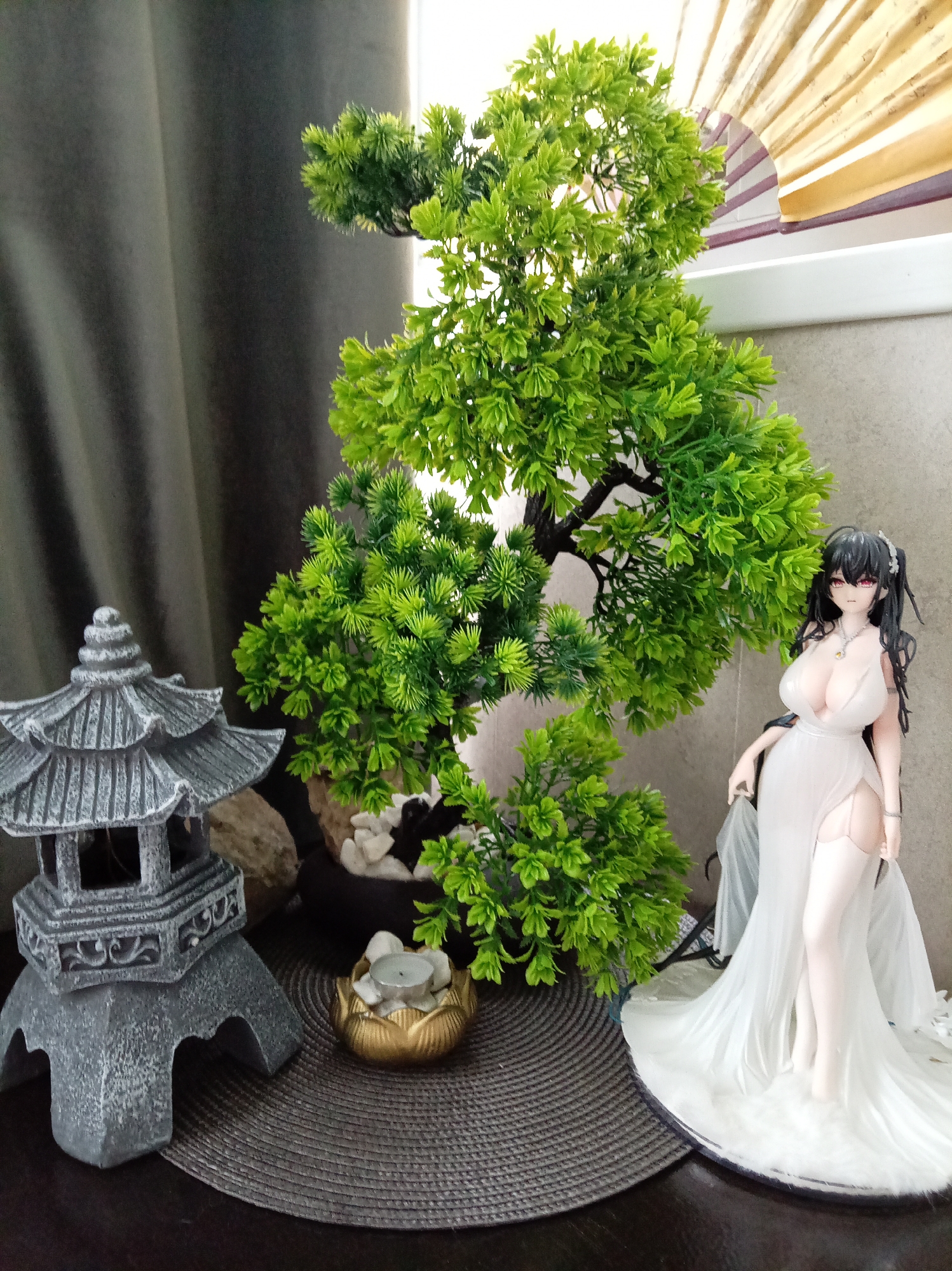 A figurine of a woman in a flowing white dress stands next to a miniature artificial bonsai tree, which is set in a pot with white stones. To the left of the bonsai tree is a small, grey, tiered pagoda ornament. In front of the bonsai tree and pagoda is a gold, lotus-shaped candle holder with a small white candle inside. The arrangement sits on a dark brown surface, possibly a table, which is covered by a grey, woven placemat. A dark grey curtain hangs in the background to the left, and a window with a partially opened blinds or fan is visible to the right, allowing light to enter the scene. The lighting suggests it could be daytime. The overall scene appears to be a decorative display, possibly for a room in a home, in Yessentuki, Russia. There is no visible text in the image.