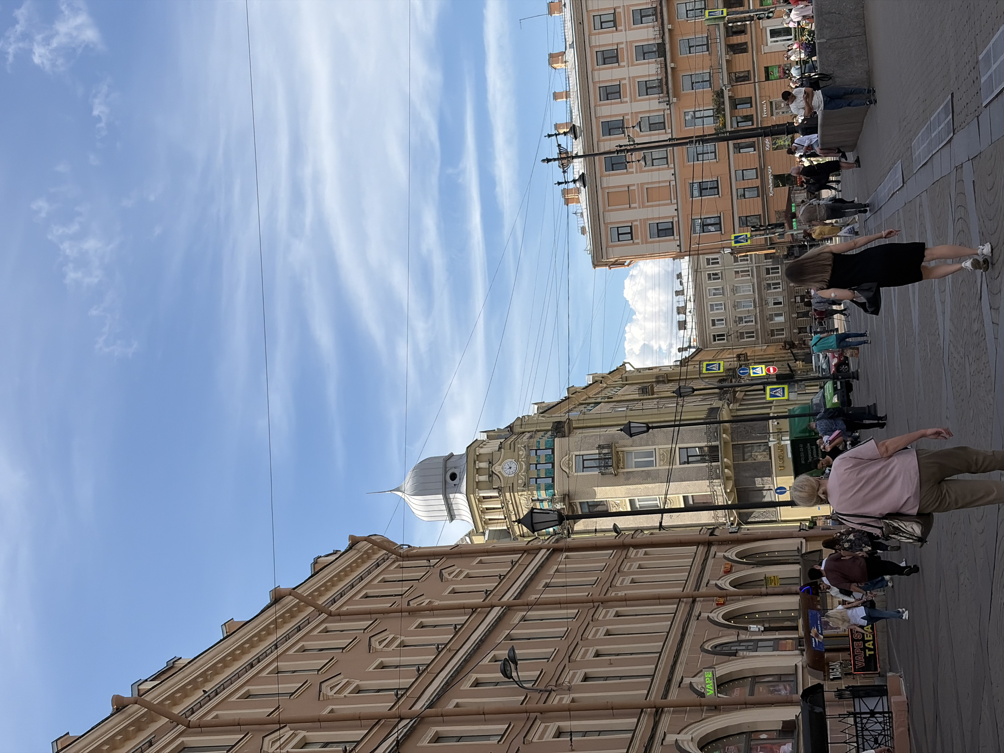 The image depicts a street scene in Saint Petersburg, Russia, on a sunny day with a partially cloudy sky. The foreground shows a paved walkway with several people walking in various directions. A woman in a black dress and white sneakers is walking away from the camera, while another woman in a pink shirt and khaki pants with a large tote bag is walking towards the camera.
The mid-ground features the facades of several buildings lining the street. The building on the left is a tall, multi-story structure with a light brown facade, arched storefronts at street level, and numerous windows on upper floors. Signs visible on this building include "VAPE" and "TABAK". Further down the street, a distinctive building with a dome-shaped roof and a clock tower is visible. Adjacent to this, an orange-colored building with numerous windows is prominent.
The background consists of more buildings and the sky. Several utility wires are strung across the scene, supported by poles. Pedestrian crossing signs and traffic signals are also present. The architecture suggests a European urban environment, consistent with Saint Petersburg.