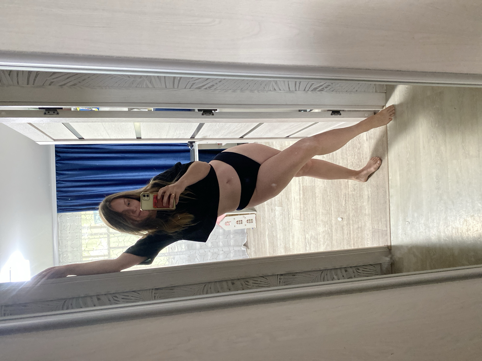 A woman is posing in front of a mirror, taking a selfie. She is wearing a black crop top and black underwear. Her left hand is on the mirror frame, and she is holding a phone in her right hand. Behind her is a partially-visible room with a blue curtained window. A small white decorative house object is on a table or surface in the background. The floor appears to be wood or laminate. The mirror is framed in white with a decorative pattern. There is no evidence to suggest that the photo was taken in Noviy, Russia.