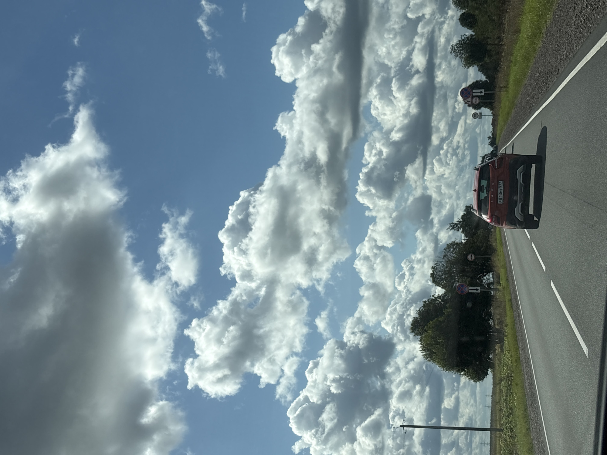 The image depicts a view from a moving vehicle on a paved road, likely in a rural or semi-rural area. The sky is partly cloudy with large cumulus formations against a bright blue sky. To the right of the road, there is a grassy shoulder and a line of trees. Road signs are visible, including one with a red circle and a diagonal red bar, and another with a number "70" inside a circle, indicating a speed limit. A red Lada automobile is traveling in the same direction in front of the viewpoint vehicle. In the distance, to the left, is an open field with what appear to be transmission towers. The specific location in the city of Voskresenskoe, Russia, is not identifiable from the visual cues in the image alone.
