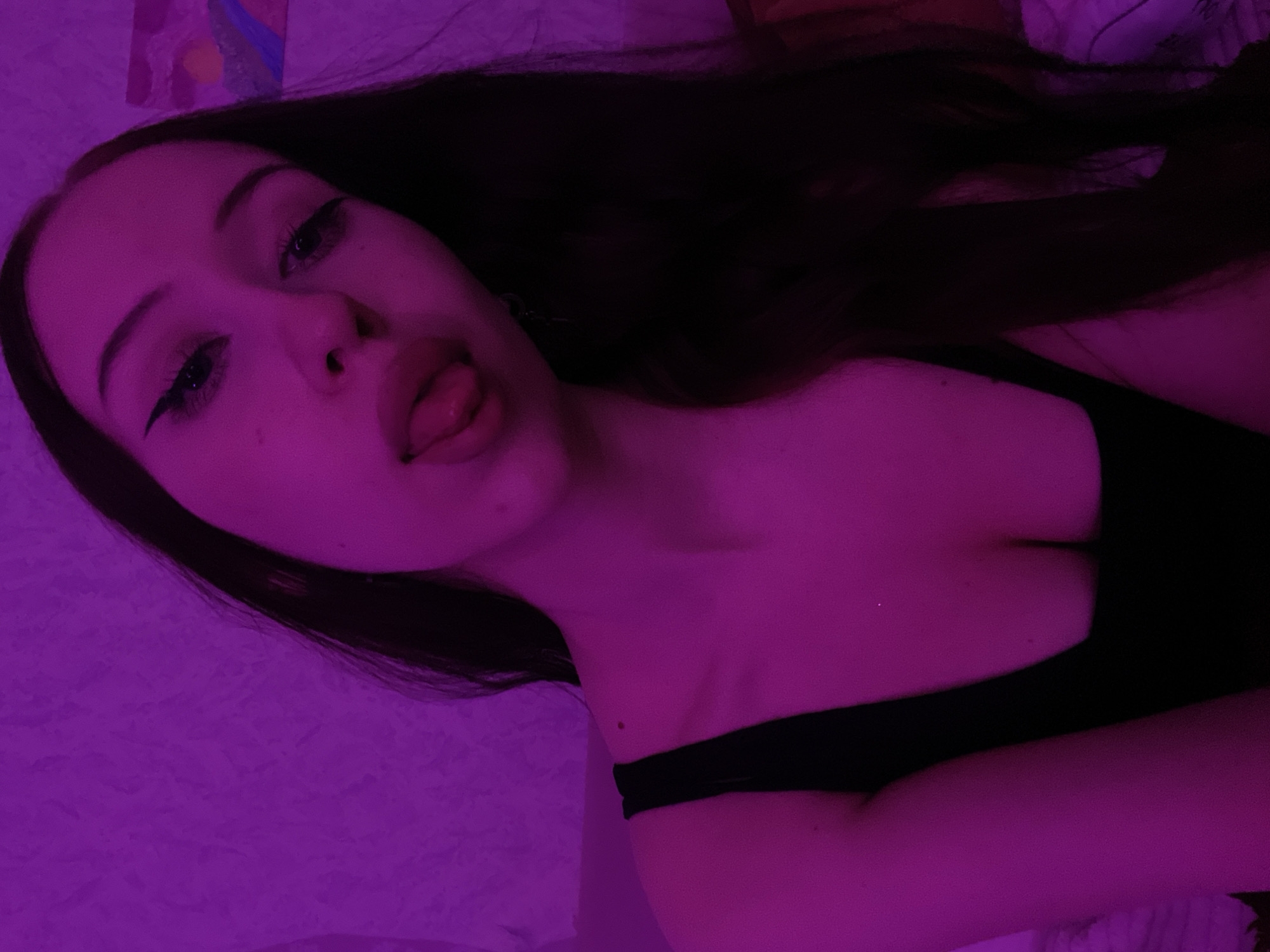 The image shows a close-up, medium shot of a young woman with long dark hair, illuminated by purple lighting. She is wearing a black top with a low neckline. Her head is tilted, and her tongue is sticking out slightly. Her makeup includes winged eyeliner. The background is a textured purple wall with a small, abstract painting in the upper right corner. There is no indication of the location within Votkinsk, Russia.