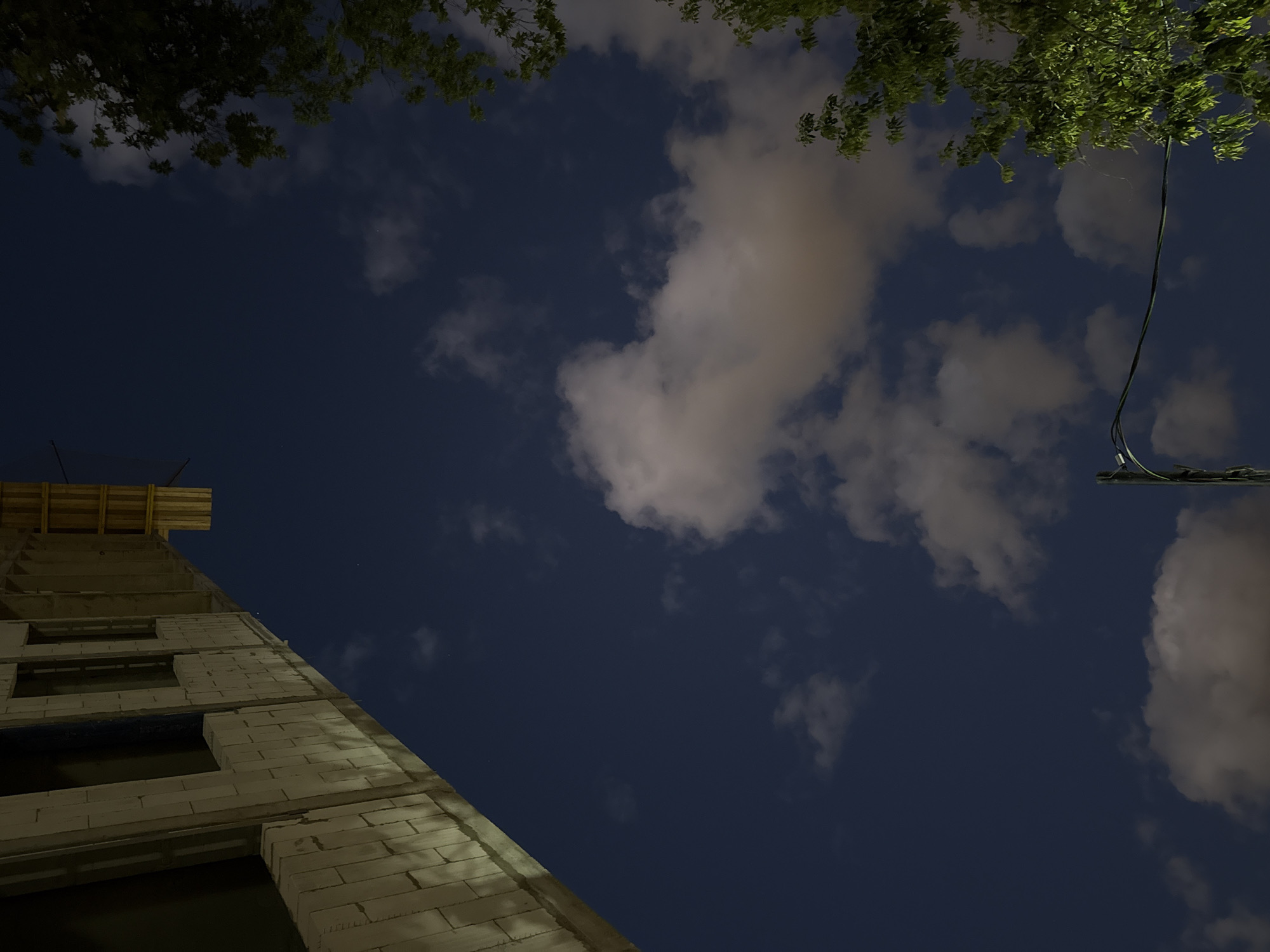 The image captures a night-time scene in Moscow, Russia, featuring the exterior of a building under construction and the night sky. The building's facade is composed of light-colored brickwork, with visible window openings. Yellow construction scaffolding is present at the top of the building. The sky is a deep blue, dotted with scattered clouds that are illuminated by an unseen light source, giving them a soft, ethereal glow. Trees with green foliage are visible on the right side of the frame, their leaves illuminated and appearing vibrant against the dark sky. A utility pole with wires is also present in the lower portion of the image, adding to the urban setting. The perspective of the shot is from below, looking upwards towards the sky and the upper sections of the building.