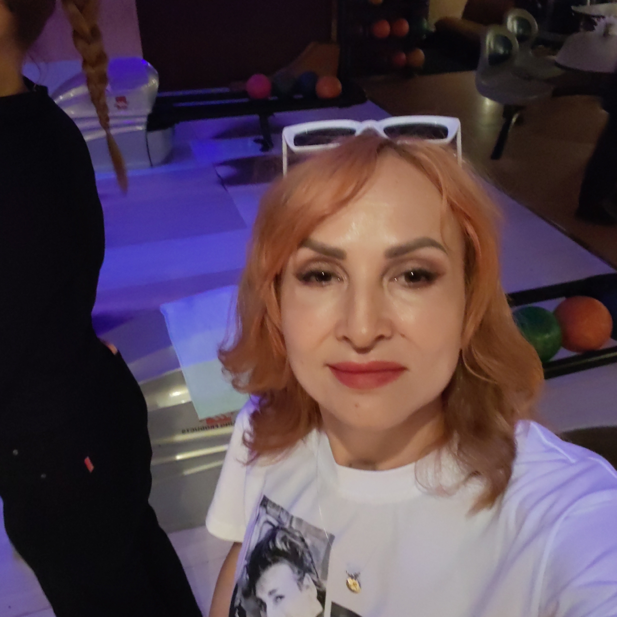 This image depicts two individuals within what appears to be a bowling alley or a similar entertainment venue in Chaikovsky, Russia, characterized by ambient purple and blue lighting.
In the foreground, a light-skinned adult female is visible from the chest up, facing the camera. She has shoulder-length, reddish-auburn hair, with white-framed sunglasses resting on top of her head. Her makeup includes eyeliner, eyeshadow, and red lipstick. She wears a white short-sleeved t-shirt featuring a black and white graphic print of a person's face on the front, along with a thin necklace holding a small gold-colored pendant. Her expression is neutral.
To the left and slightly behind the foreground subject, the left side and back of a second individual are visible. This person has long, light-colored braided hair extending down their back and is wearing dark, possibly black, clothing, including what appears to be black pants with a small red tag on the back pocket area. This individual's back is turned towards the camera.
The environment includes elements typical of a bowling alley. The floor is illuminated with a strong purple-blue hue. On the right side, a bowling ball return system is visible, holding a green and an orange bowling ball. In the deeper background, a rack contains several orange and dark-colored bowling balls. Further into the background on the right, a dark wooden table and a metallic chair with a distinctive, possibly stylized design are present. On the left side of the frame, behind the second person, a seating area or bench is partially visible. A light-colored piece of paper or signage rests on a surface in the mid-ground.