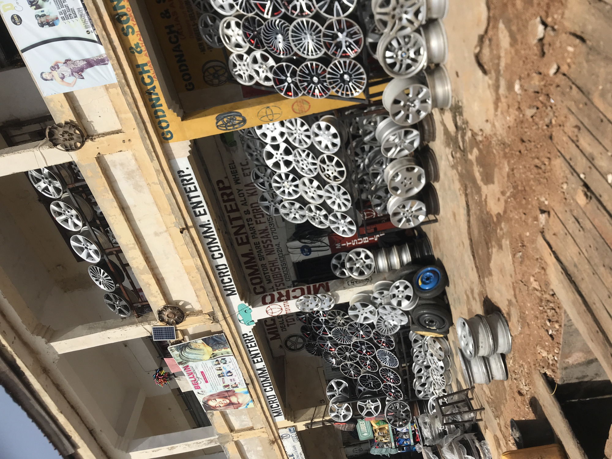 The image captures a commercial street scene, likely a market in New Stadium, Nigeria, under bright daylight. The primary subjects are numerous car wheel rims, predominantly silver and grey alloy wheels, displayed for sale. They are stacked in rows, both horizontally and vertically, with a wide variety of designs and sizes, many appearing new. Older, possibly used, car hubcaps are also visible.
Two main businesses are identified by prominent signage. "MICRO COMM. ENTERP." is visible multiple times, advertising "LL MOTOR SPARE PARTS & ALOY WHEEL" and specifying brands like "MITSUBISHI, NISSAN, FORD KIA ETC." They also deal in "TOYOTA." Another business, "GODNACH & SON (ALIAS ALHAJI YARO)," advertises "NEW WHEELS - BELGIUM." An advertisement for "LUBREX" is also seen.
Adjacent to the wheel shops, a large banner for "BELLVIN Signature" advertises fashion and beauty services, including "ASO EBI DRESSES," "BESPOKE DRESS," "RTW (READY TO WEAR)," and "MAKEUP," featuring images of women. A small solar panel is affixed to a building structure. The ground level is a mix of dirt and concrete, typical of an outdoor market setting. Bright sunlight indicates a clear, sunny day. Phone numbers (08162786784, 09035100214, 0703154) are present on various banners for contact.