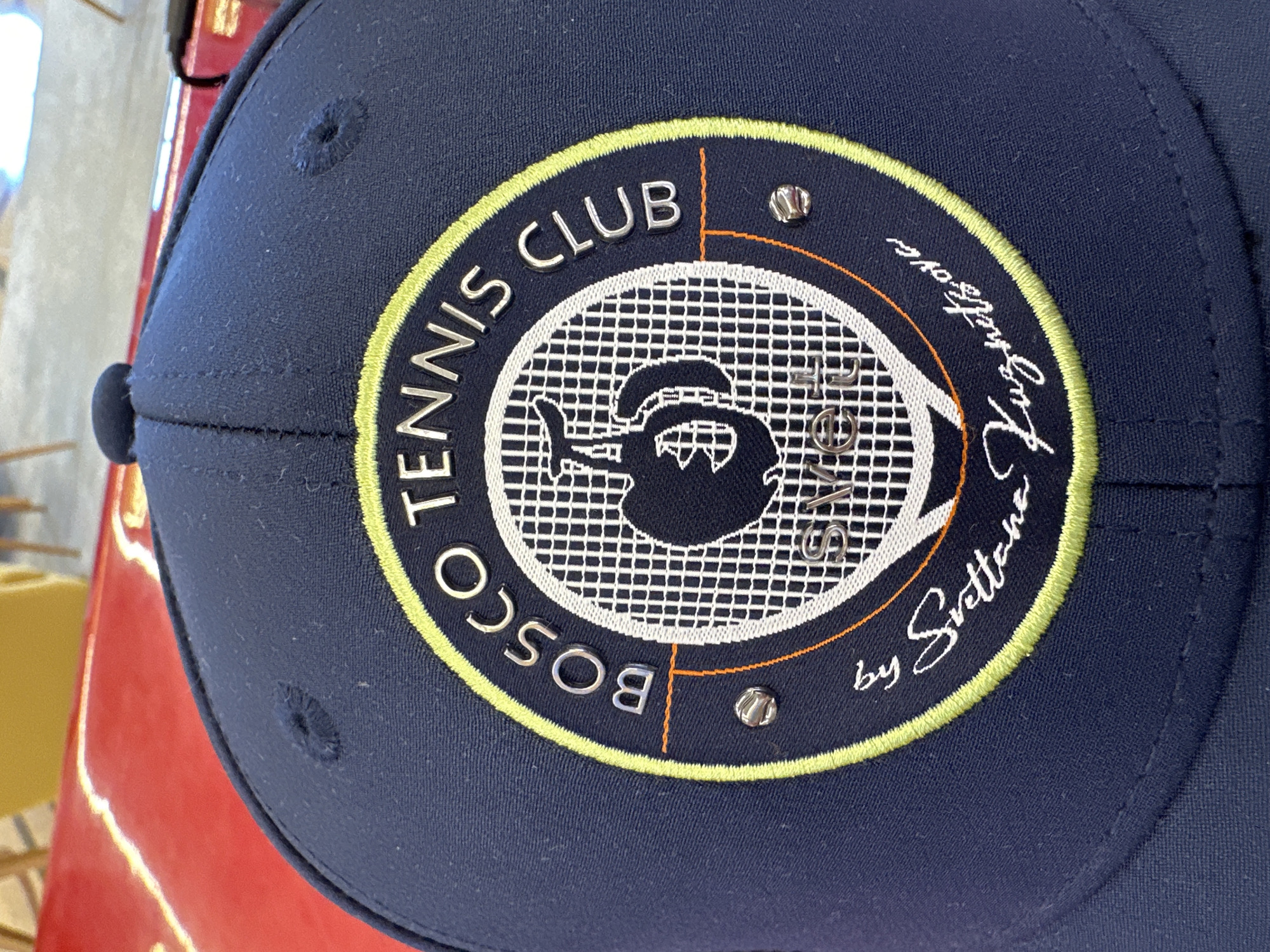 The image displays a close-up of a navy blue baseball cap, likely positioned indoors on a reflective red surface, possibly a display shelf or table. The cap features a prominent circular embroidered emblem on its front.
The emblem's design incorporates a tennis theme. Around the top and left arc of the circle, the words "BOSCO TENNIS CLUB" are spelled out in raised silver-colored letters against the dark blue background of the emblem. A light green/yellow embroidered border outlines the main circle.
Within the circle, a white tennis racket head with visible strings is depicted. Centered on the racket strings is a stylized dark blue silhouette of a teapot or kettle. On the right side of the emblem, following the curve, is text "by Svetlana Lungart" (the last word being somewhat obscured but appears to be "Lungart"). Vertically positioned on the right side within the racket strings, the word "SVETLA" is visible. Two small, decorative silver studs are placed on the emblem, one near "CLUB" and another near "by Svetlana". Additional thin orange lines form a partial inner border and accents within the emblem.
The cap itself appears to be made of a textured fabric. The background behind the cap is softly blurred, showing hints of light-colored objects and surfaces, further suggesting an indoor setting. The lighting is artificial, causing reflections on the red surface. Given the "BOSCO" branding and the provided context, the item is associated with Sochi, Russia.