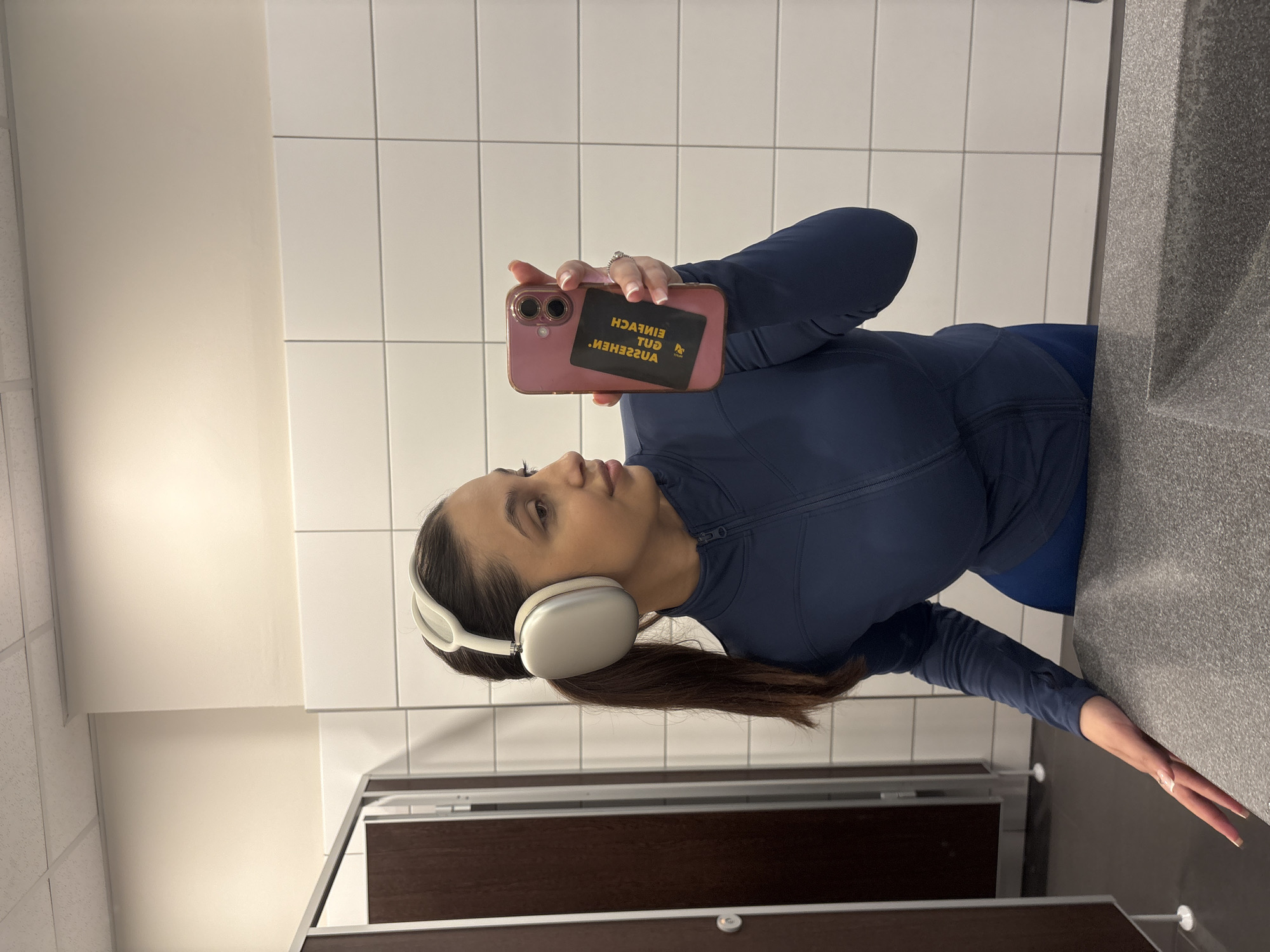 A woman is taking a selfie in a bathroom. She is wearing a blue athletic jacket and white headphones. Her hair is pulled back in a ponytail. She is holding a pink phone with a black card attached to the back. The background consists of white tiles and a toilet stall. The location is in Mönchengladbach, Germany.