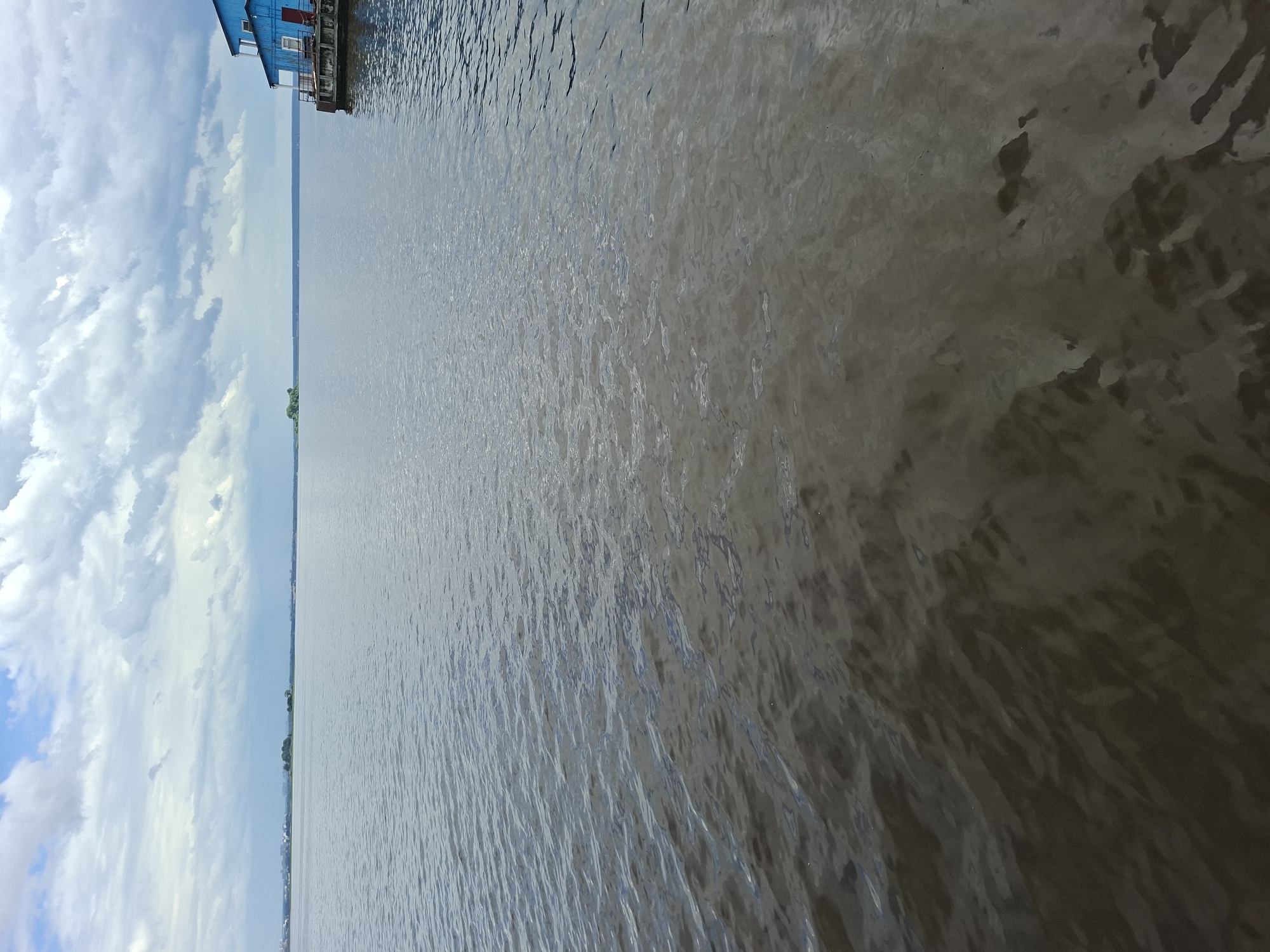 This is a landscape photograph taken from a dock or pier, featuring a view of a large body of water under a cloudy sky. On the left side of the image, a section of a bright blue building with red accents is visible, suggesting it is a structure on the shore or a boathouse. The building has windows and a railing, indicating it's accessible.
The majority of the frame is dominated by the water, which appears somewhat murky or silted, with ripples and light reflections creating texture across its surface. The water extends to the horizon, where it meets a hazy outline of distant land, possibly featuring trees and some structures, characteristic of a shoreline. The sky is filled with dramatic, white and grey clouds, with patches of blue sky peeking through, suggesting either an overcast day with breaks of sun or a time of day when the clouds are particularly active.
The scene evokes a sense of calm and perhaps a touch of melancholy due to the cloudy sky. The location context of Verhnij Uslon, Russia, implies a riverside or lakeside setting, common for such structures. The time of day is not explicitly clear but likely daytime due to the brightness of the sky and the reflections on the water. There is no visible text in the image.