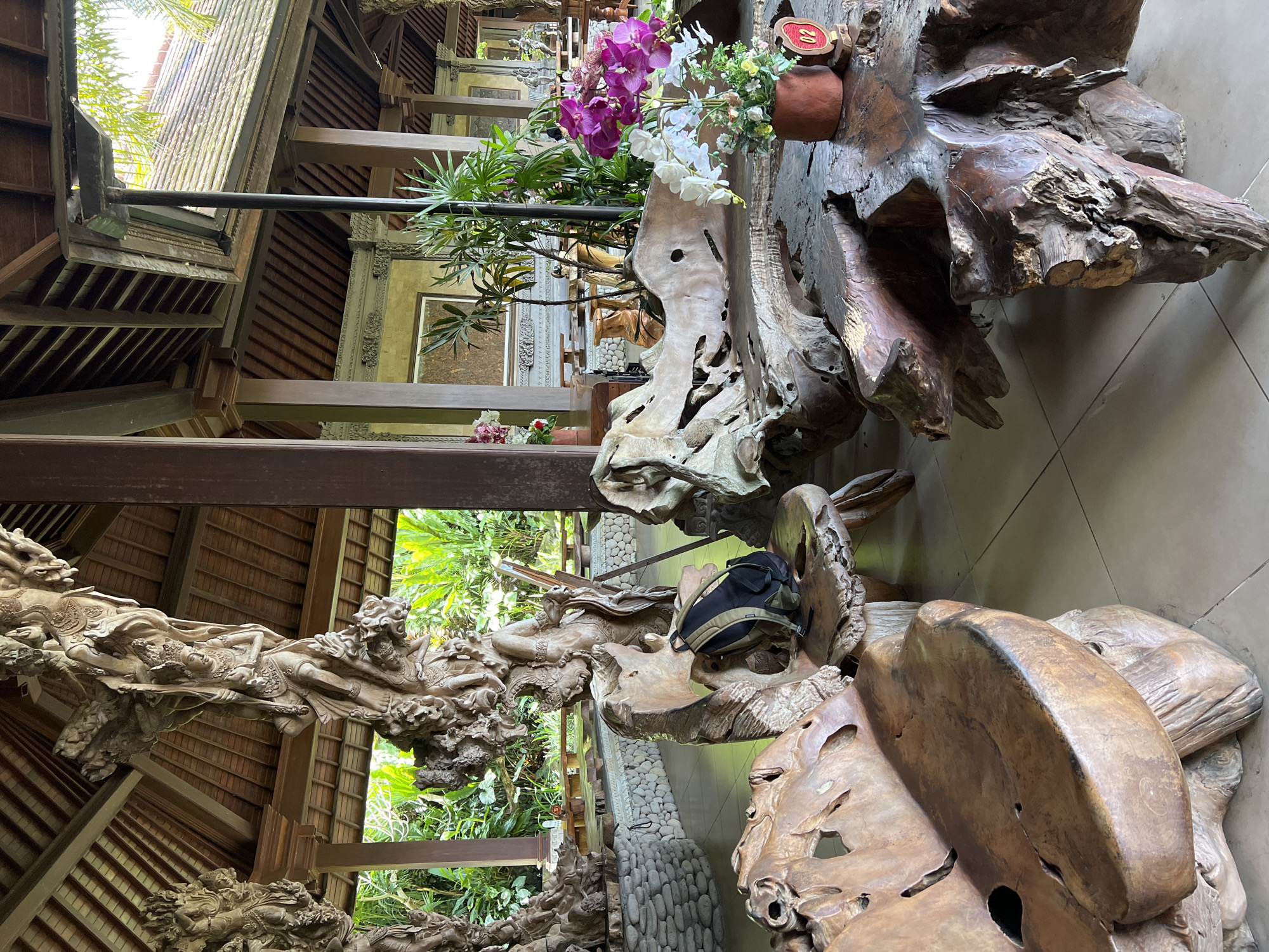 The image depicts an interior space in Kedewatan, Indonesia. A large, intricately carved wooden structure dominates the scene, featuring figures and details. The structure appears to be part of a building or a decorative element within it. The setting includes a tiled floor. The space is decorated with flowers and plants. A black backpack sits on top of the wooden structure. The architecture includes detailed carvings and wooden beams.
