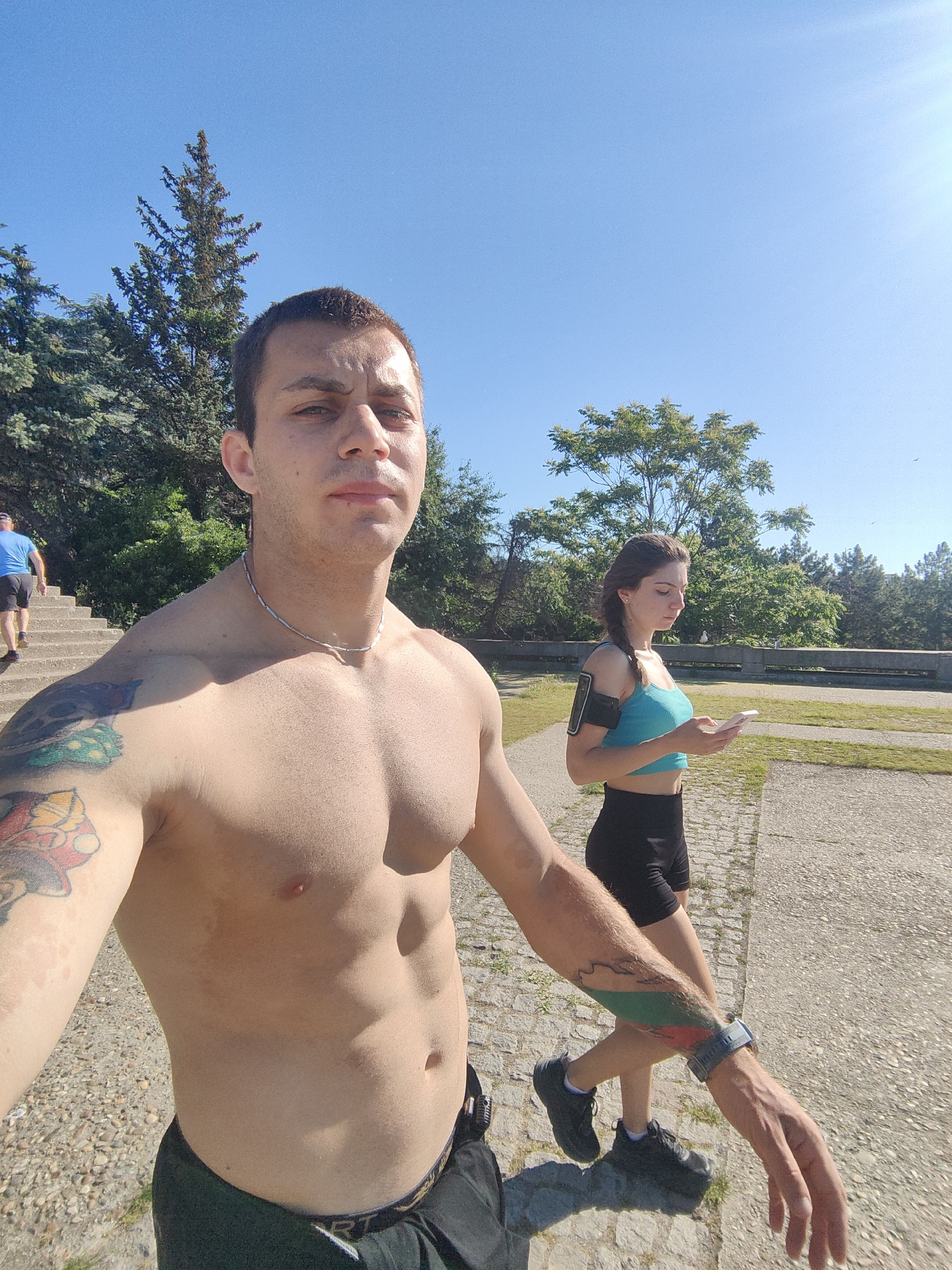 A man is taking a selfie in front of a park with a woman in the background. The man is shirtless and wearing black shorts and a necklace. He has tattoos on his arms. The woman is wearing a blue top and black shorts, with an arm band on her left arm. She is holding a phone and looking down at it. Another man in a blue shirt is walking up stairs in the background. The scene is outdoors on a sunny day, with trees and a clear blue sky. The ground is paved with stone.