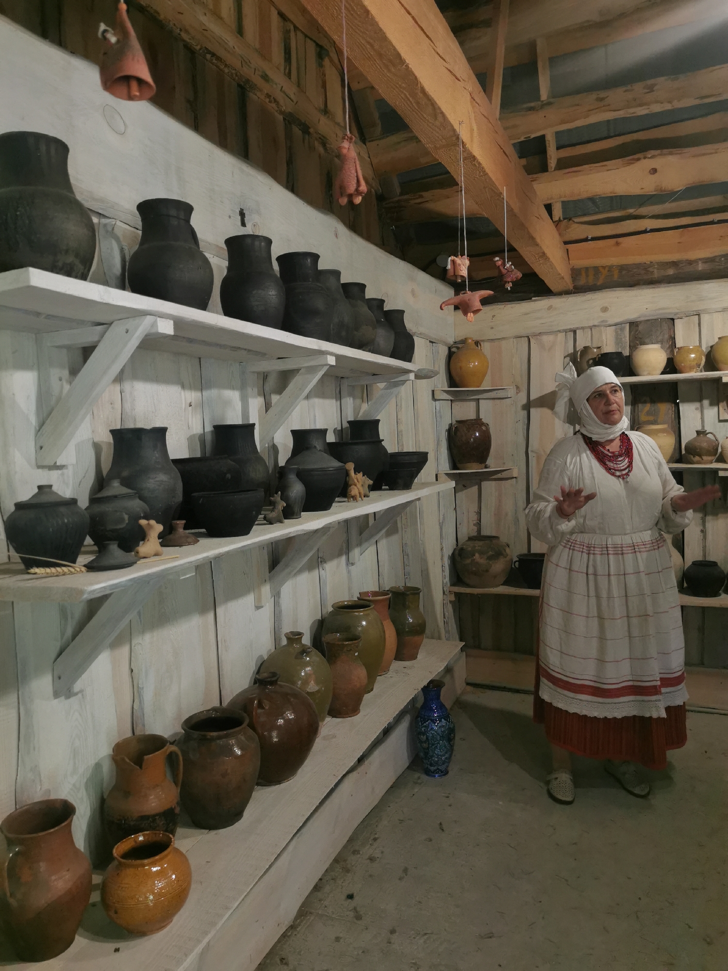 A woman dressed in traditional Belarusian clothing stands in a rustic, wooden-walled room filled with pottery. The room is likely a museum or workshop, given the display of various clay vessels on shelves lining the walls. The woman, wearing a white headscarf, a white blouse with embroidered trim, and a red and white striped skirt, gestures with her hands as if explaining something. The pottery includes numerous dark, unglazed pots and pitchers, as well as some glazed earthenware in earth tones. Decorative clay figures hang from the ceiling beams. The floor is concrete, and the overall impression is one of a historical or cultural exhibit. The specific location is Шчарчоўскі сельскі Савет, Belarus.