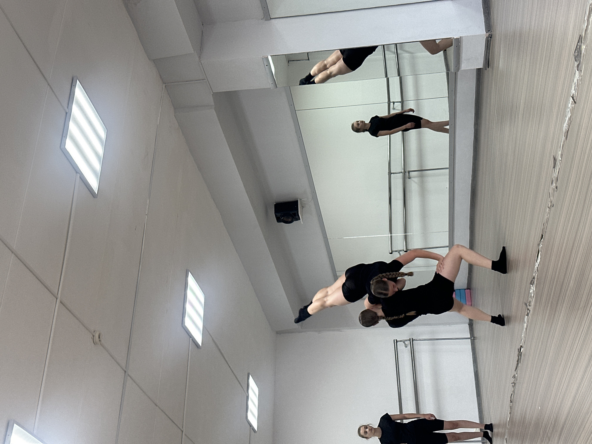 In a dance studio, two young women in black athletic attire are engaged in an acrobatic dance move. One woman is being held aloft by the other, with her body inverted and legs extended. A third young woman stands to the left, observing the performance. The studio features a polished wooden floor, a barre along the wall, and large mirrors that reflect the scene. Fluorescent lighting panels are mounted on the ceiling, and a smoke detector is visible. The overall environment suggests a training or rehearsal space for dance. There is no information within the image that indicates a specific location within Votkinsk, Russia.