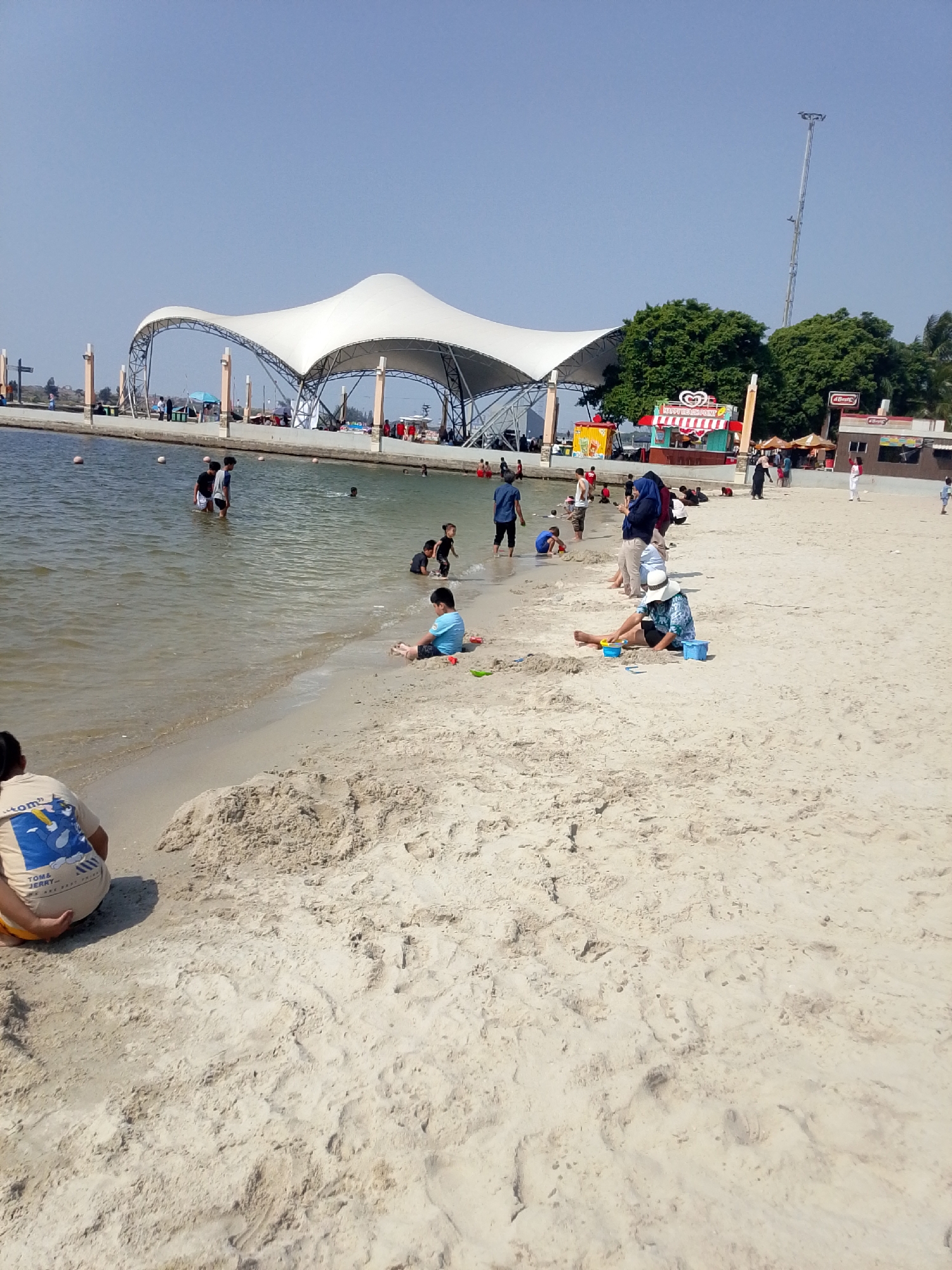 The image shows a bright, sunny day at a beach resort, likely Ancol Beach City in North Jakarta, Indonesia. A large, white, tent-like structure with a curved, geodesic dome roof is prominent in the background, adjacent to a body of water. Several people are visible on the beach and in the shallow water. On the sand in the foreground, a person is sitting and playing with sand toys, with two blue buckets and a yellow spade. Further down the beach, more people are seen wading in the water and playing. There are also some small food stalls and other structures visible along the waterfront. The sand is light-colored and appears to be a popular recreation area. The sky is clear and blue, suggesting a pleasant day for outdoor activities.