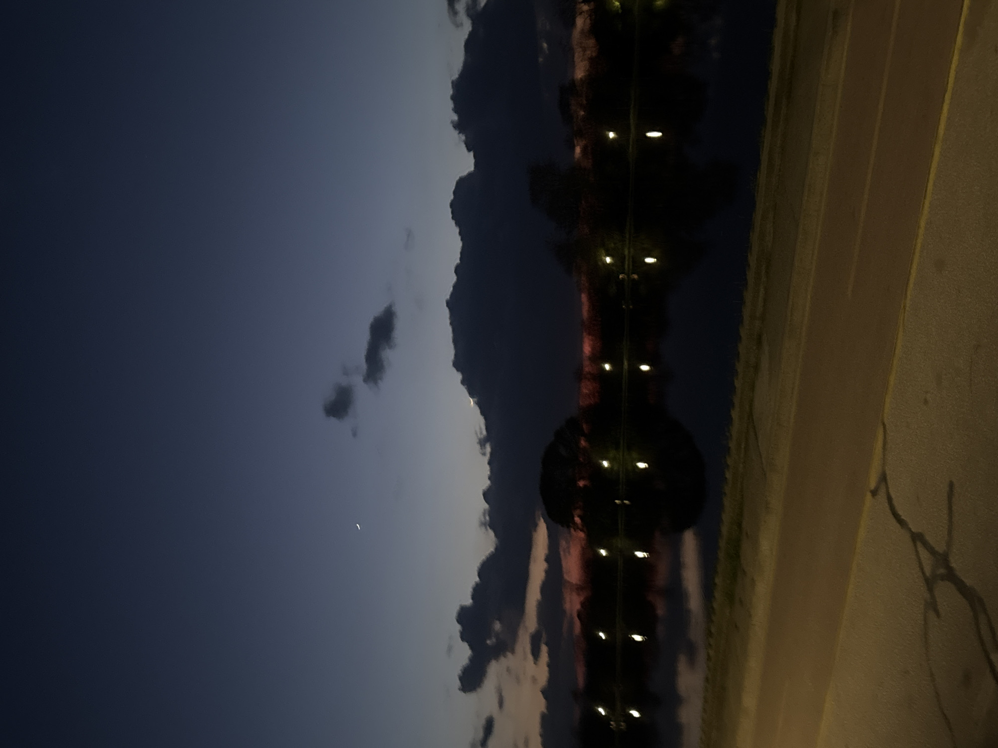 The image captures a nighttime scene in Plovdiv, Bulgaria. The sky, transitioning from a deep blue to lighter shades, features scattered clouds and a faint crescent moon. A row of lights reflects on the water surface near trees and a roadway. The roadway, illuminated by artificial light, shows some surface cracking. No people are visible.