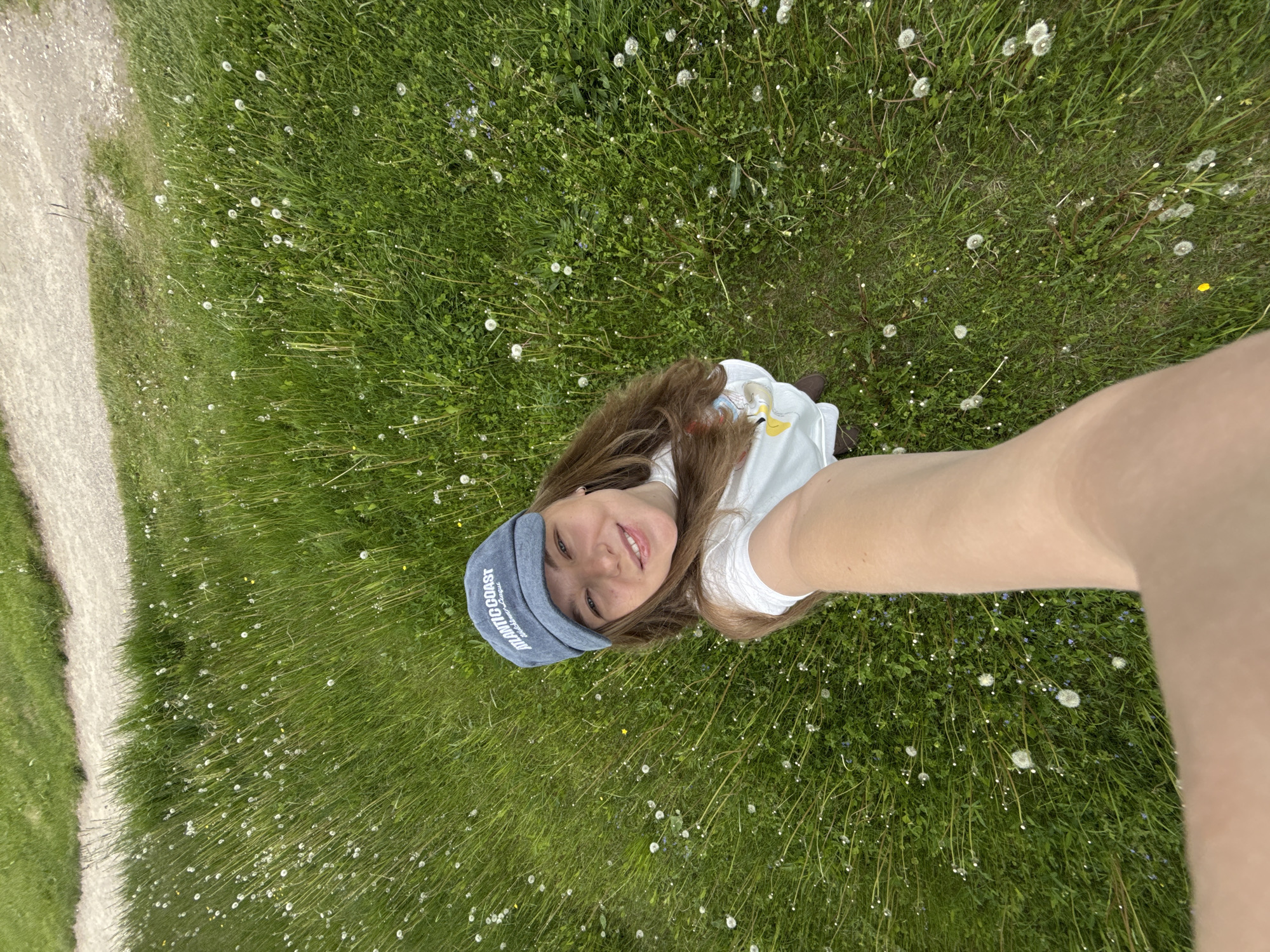 A cheerful young woman is captured in a selfie, taken from an overhead perspective as she lies or reclines in a vibrant green field. She has long brown hair and is smiling pleasantly at the camera. She wears a white t-shirt and a light blue, denim-style cap with "ATLANTIC COAST" clearly visible in white lettering on the brim. Hints of brown shoes are visible near her feet.
The setting is a lush natural landscape, characterized by abundant green grass densely populated with countless white dandelion seed heads. There are also scattered small blue flowers and other wild plants within the foliage. A light-colored dirt or gravel path runs along the left side of the frame, winding through the natural environment. The scene is bathed in bright, natural daylight, indicating a clear day. The overall impression is one of relaxation and enjoyment in nature. The location is Dobrograd, Russia.