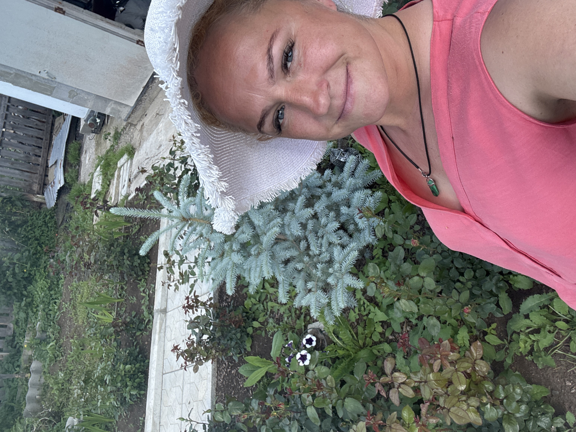 A woman with blonde hair and blue eyes, wearing a white wide-brimmed hat and a pink top, smiles at the camera. She is wearing a dark necklace with a green pendant. She is outdoors, in what appears to be a garden with various plants and a small blue spruce tree. In the background, a wooden fence and some construction materials are visible. The scene is likely located in Chaikovsky, Russia.