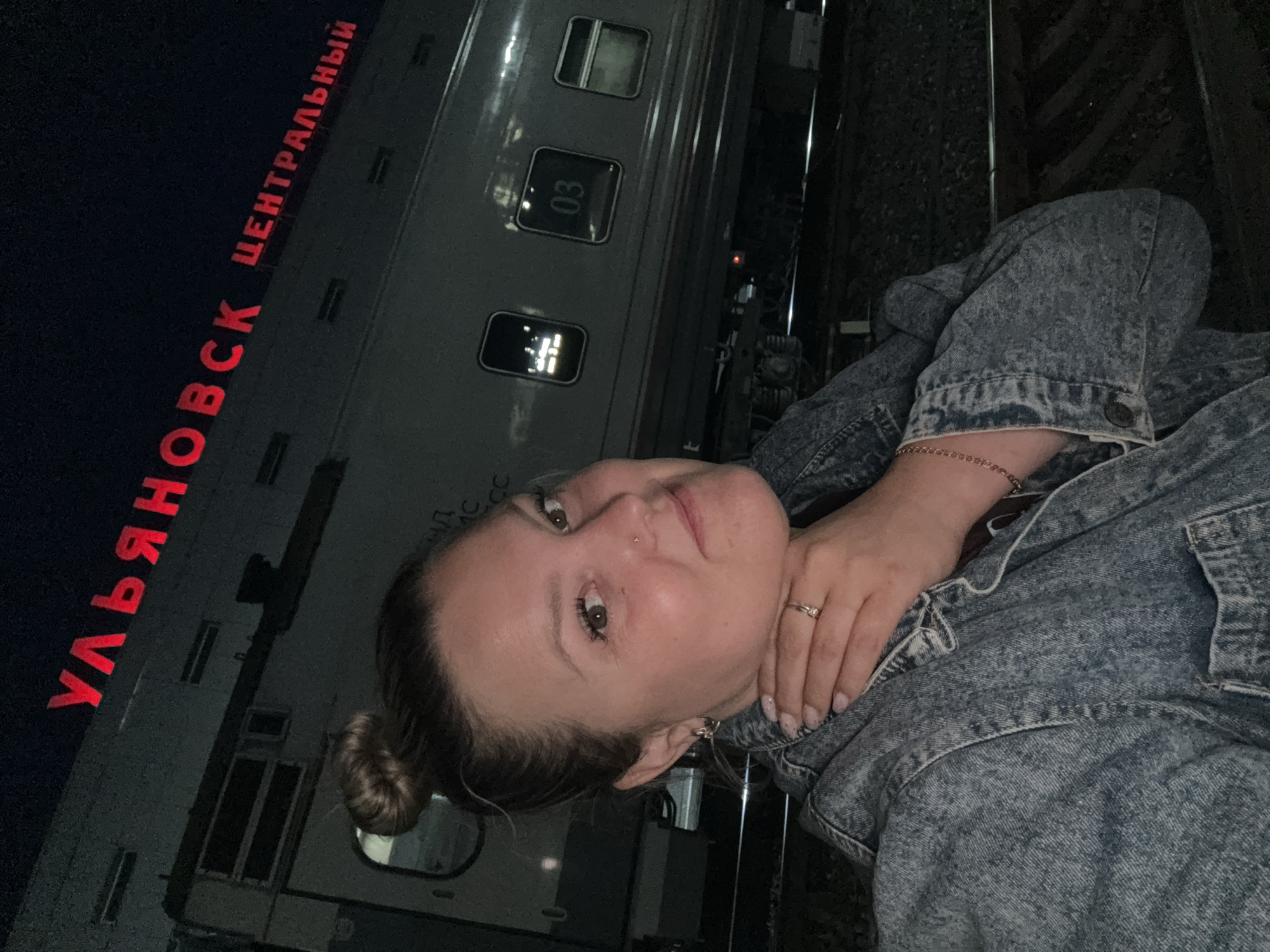 A woman with her hair in a bun, wearing a denim jacket, is taking a selfie at night in front of a train. The train car visible has a window with the number "03" displayed. Above the train, red illuminated Russian lettering reads "УЛЬЯНОВСК ЦЕНТРАЛЬНЫЙ" (Ulyanovsk Central). The setting appears to be a train station in Ulyanovsk, Russia, with train tracks visible in the foreground.