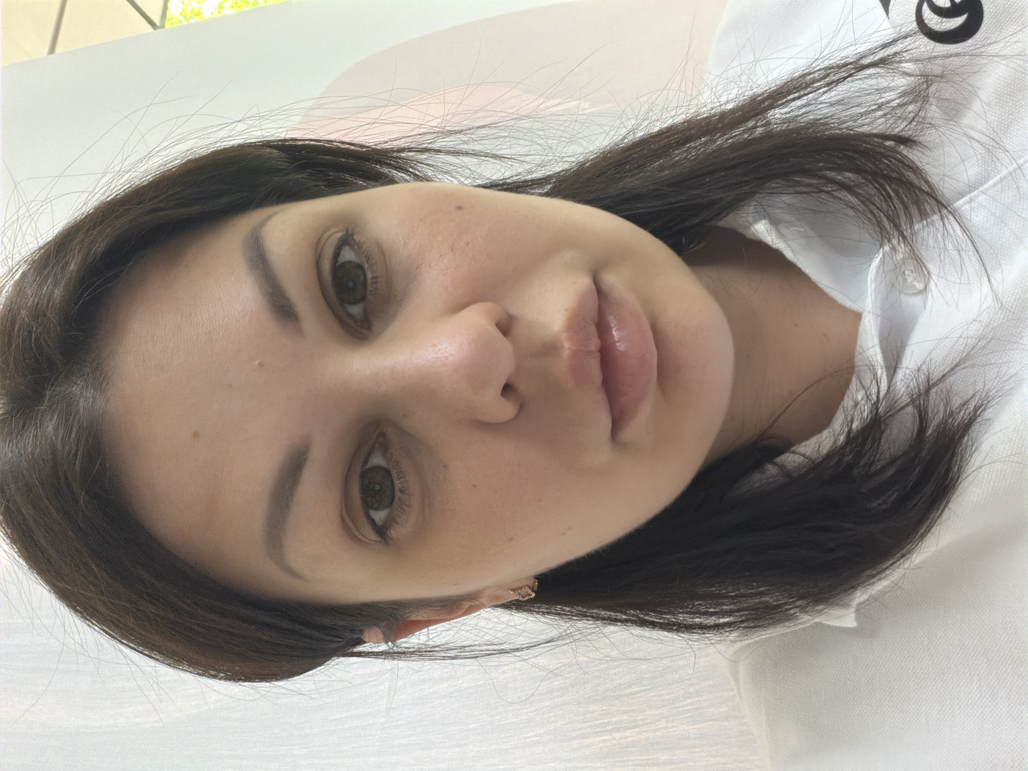 The image captures a close-up portrait of a woman in Sochi, Russia. The subject is a female, appearing to be in her late 20s or early 30s, with dark brown hair parted on her left side and falling loosely around her face and shoulders. She has a light olive complexion, brown eyes, and maintains a neutral expression while looking directly at the camera. Her eyebrows are well-defined, and her lips appear full with a subtle gloss. A small, dark mole is visible on her right cheek (viewer's left), below the outer corner of her eye. A small, golden-colored earring, possibly an intricate design, is worn in her left ear (viewer's right). She is dressed in a white collared shirt, likely a polo, featuring a pique texture. A single clear button is visible at the neckline, and a partial black logo, shaped like a stylized 'C' or similar curve, is visible on the viewer's left side of the shirt. The background is out of focus; the right side (viewer's right) shows a light-colored, vertically textured surface, while the left side (viewer's left) reveals soft, light pink or peach tones with vertical elements, and a hint of green foliage and bright outdoor light in the upper left corner, suggesting an interior location near a window or open area.