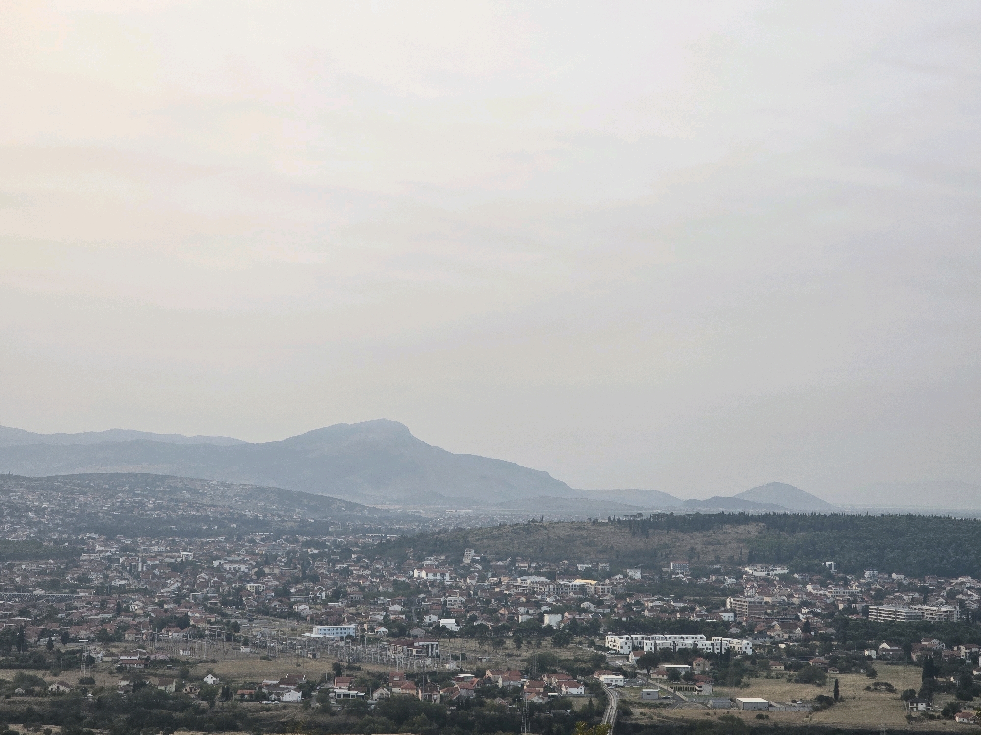 This is a wide, elevated, over-the-shoulder, landscape shot of Podgorica, Montenegro, under a hazy sky. The camera is positioned in the foreground looking down and out over a sprawling residential area. The city appears densely packed with many buildings, most of which are small, two-story homes with red-tiled roofs. There are also larger, more modern buildings scattered throughout. In the distance, rolling hills and mountains rise up, also appearing hazy. The overall scene is muted, with soft colors, and the atmospheric perspective creates a sense of depth and distance. The lighting is overcast, suggesting a cloudy or hazy day. There are no visible people in the image.